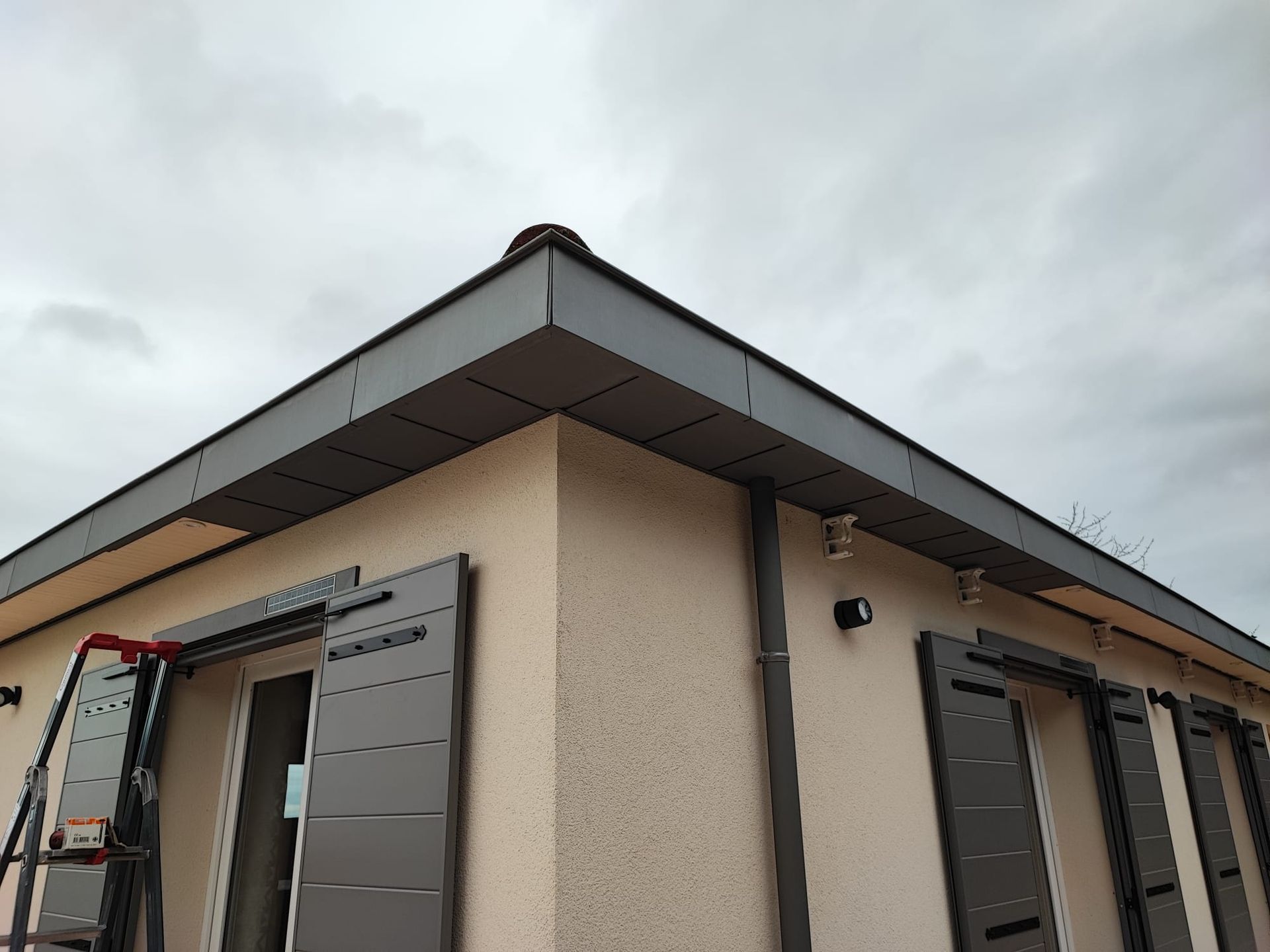 Coin d'une maison en stuc beige avec des boiseries grises et un toit sombre.