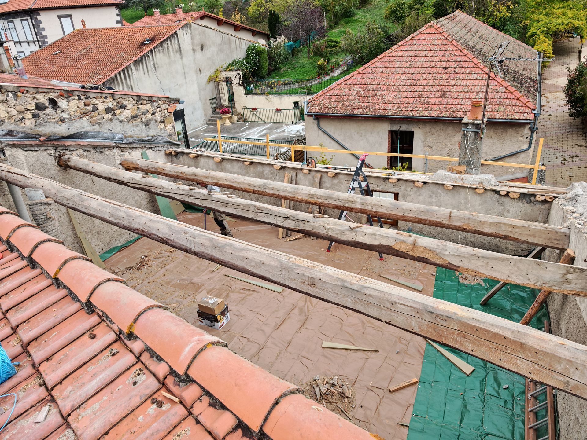 Chantier de construction sur un toit avec poutres en bois, toits de tuiles rouges et maisons délabrées en arrière-plan.