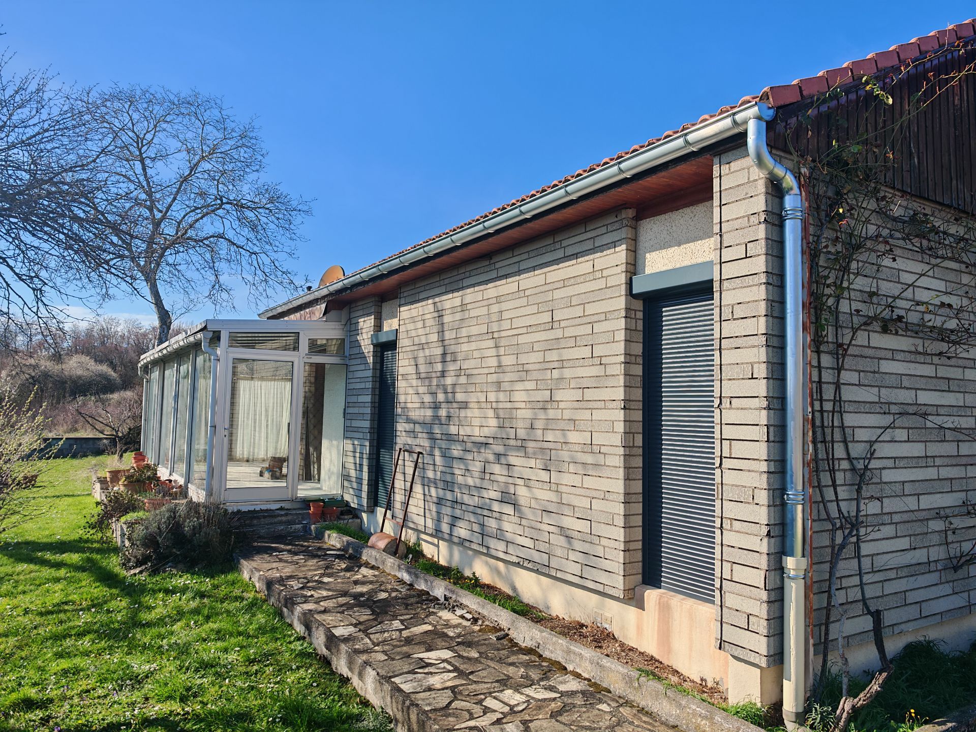 Vue latérale d'une maison en briques avec une véranda, un ciel bleu et une cour gazonnée avec un chemin de pierre.