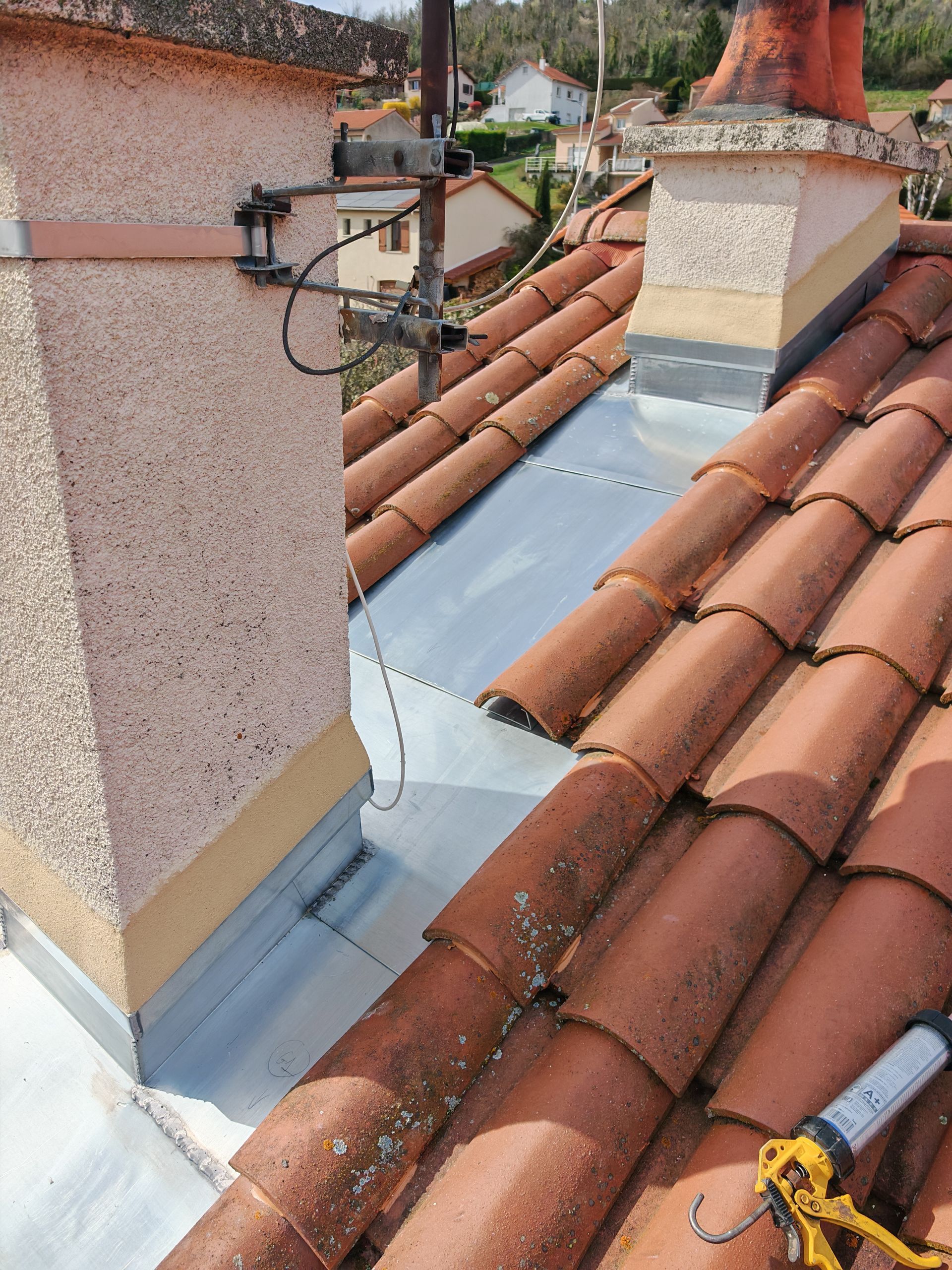 Cheminée de toit et solins argentés entre les toits de tuiles rouges et une antenne de télévision à proximité.