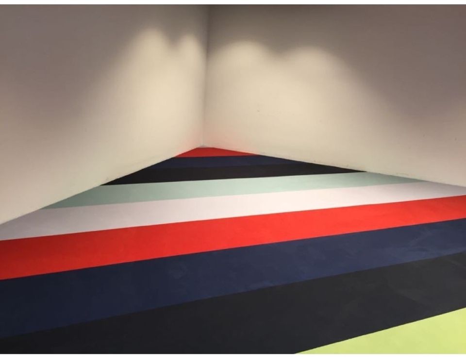 Zimmerecke mit gestreiftem Fussboden in Rot, Blau, Grün und Grau. Weisse Wände.