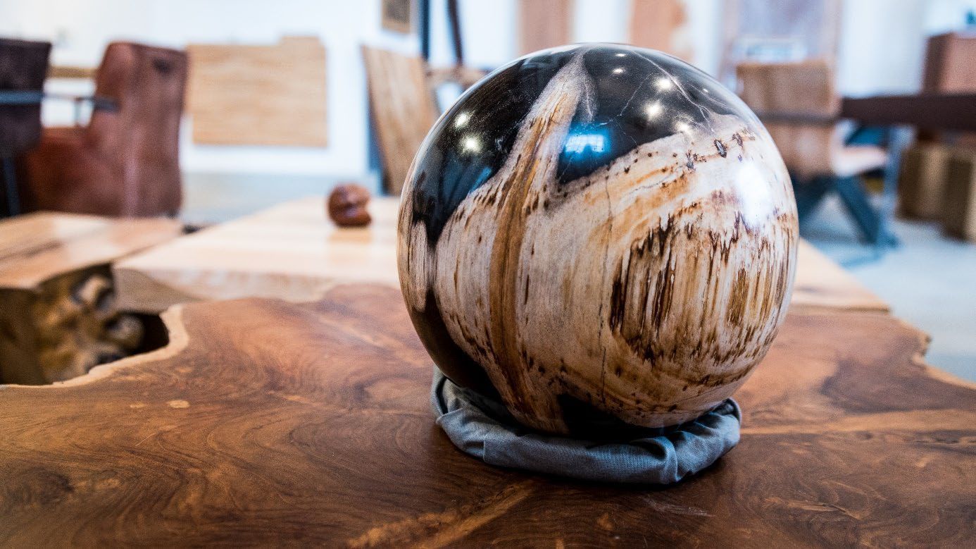 Glänzende Kugel mit Holzmaserung liegt auf einem dunklen Stoffpolster auf einer rustikalen Holzplatte.