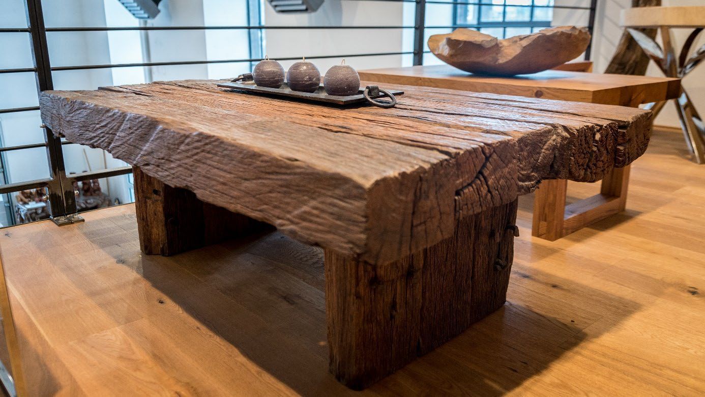 Massiver, rustikaler Holztisch mit unregelmäßiger, naturbelassener Oberfläche und dicken Beinen in einem hellen Raum.