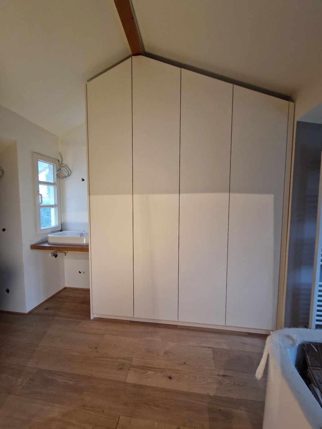 Schrank mit vier weißen Türen in einer Ecke eines Raums mit Holzboden und Waschbecken an der linken Wand.