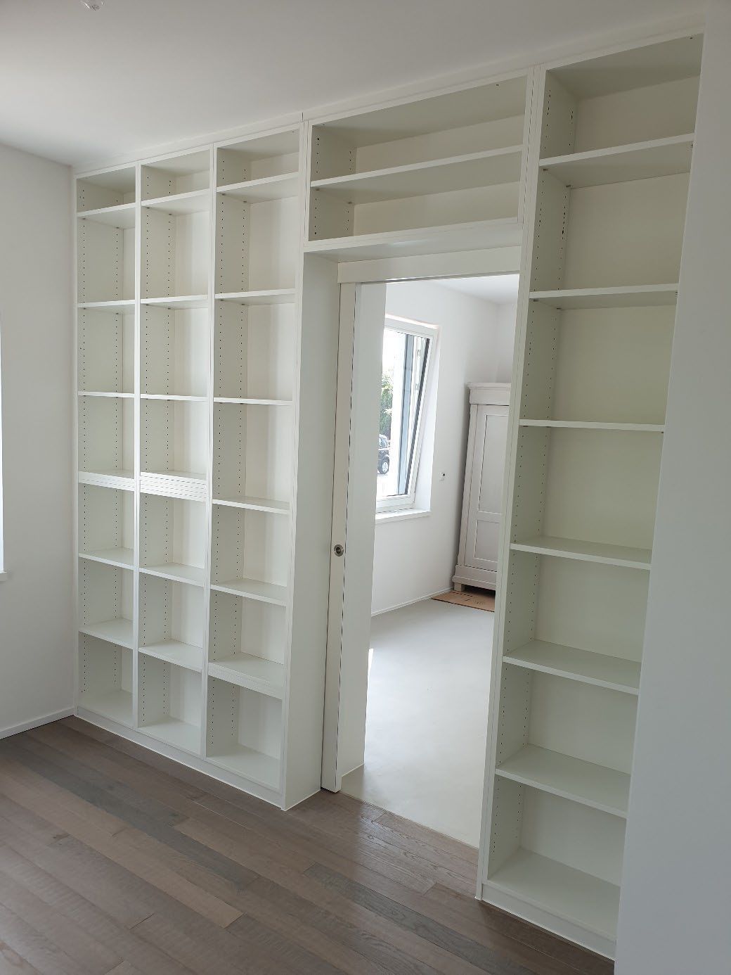 Weiße Einbauregale mit mehreren Fächern umrahmen eine offene Tür in einem hellen Raum mit Holzboden.