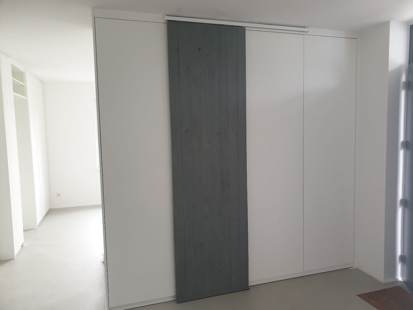 Weißer Einbauschrank mit einer grauen Schiebetür in einem hellen Raum mit weißem Boden.