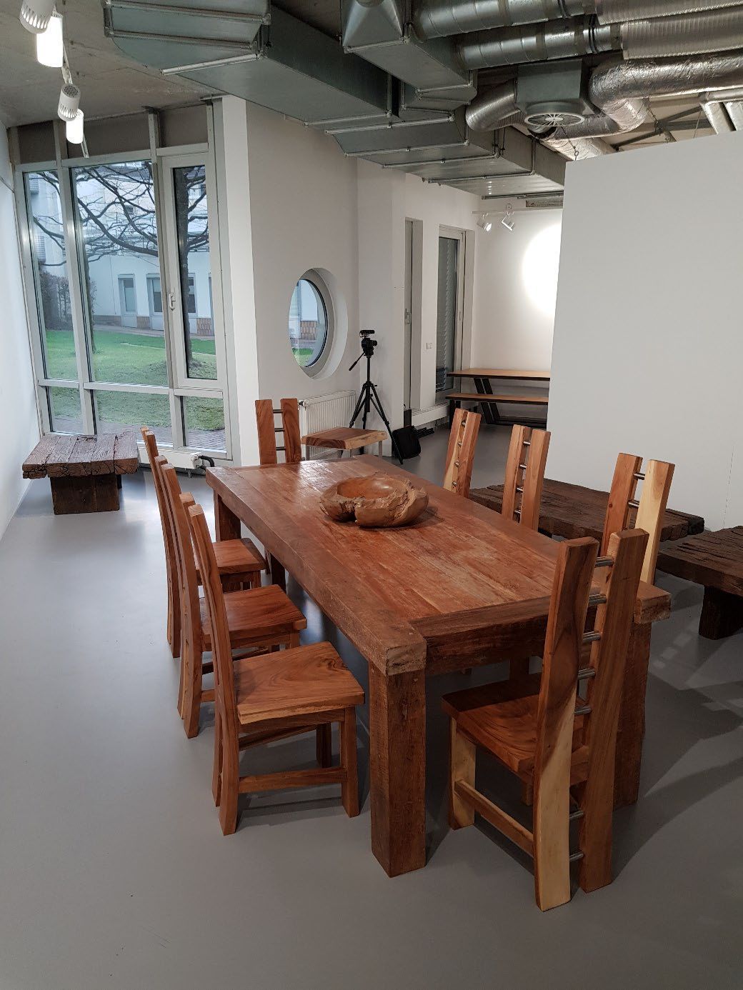 Holztisch mit sechs Stühlen in einem hellen Raum mit großen Fenstern und sichtbaren Rohrleitungen an der Decke.