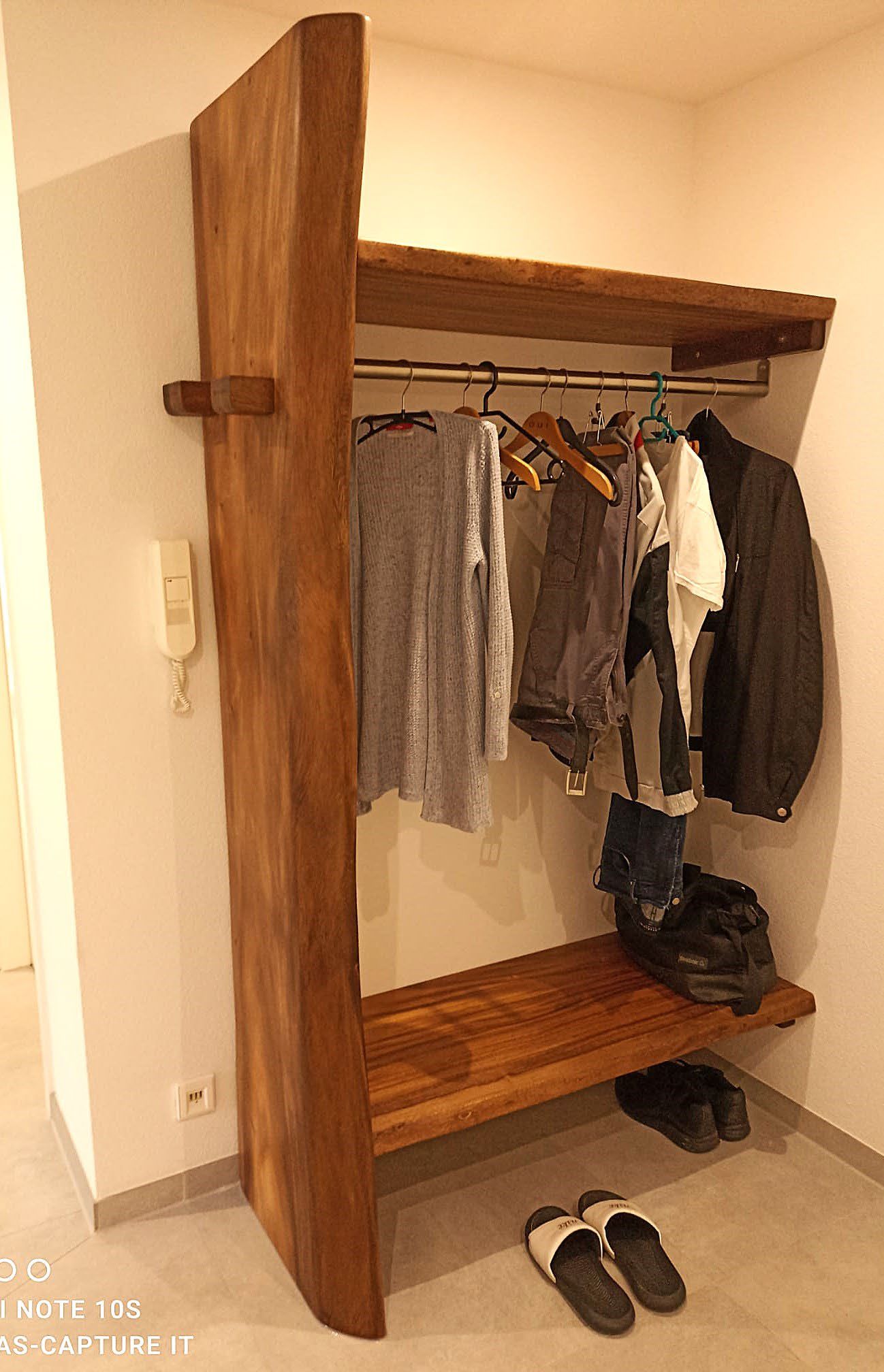 Offener Garderobenschrank aus Holz mit mehreren Kleidungsstücken an Kleiderbügeln und Schuhen darunter.