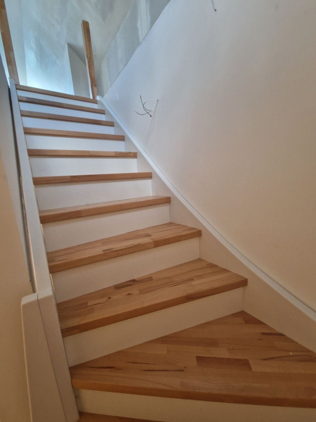 Holztreppe mit weißen Setzstufen und weißem Geländer an der linken Seite, rechts weiße Wand mit sichtbaren Kabeln.