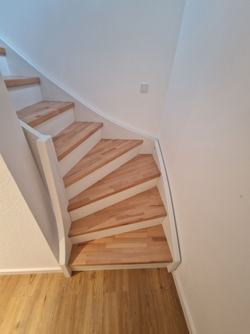 Holztreppe mit gewendelten Stufen und weißem Geländer in einem hellen Innenraum.