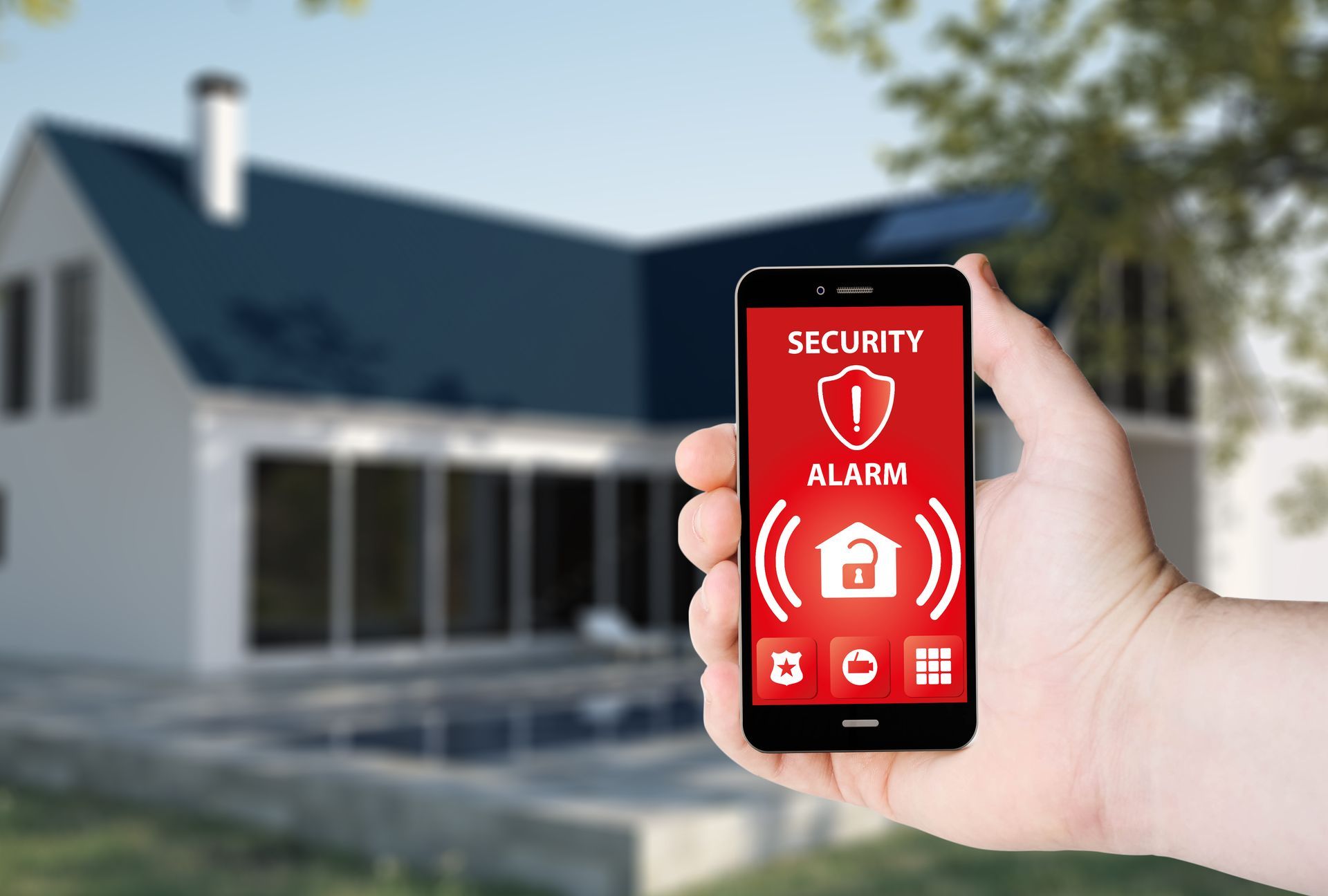 Une main tenant un smartphone avec un écran rouge d'alarme de sécurité domestique devant une maison floue