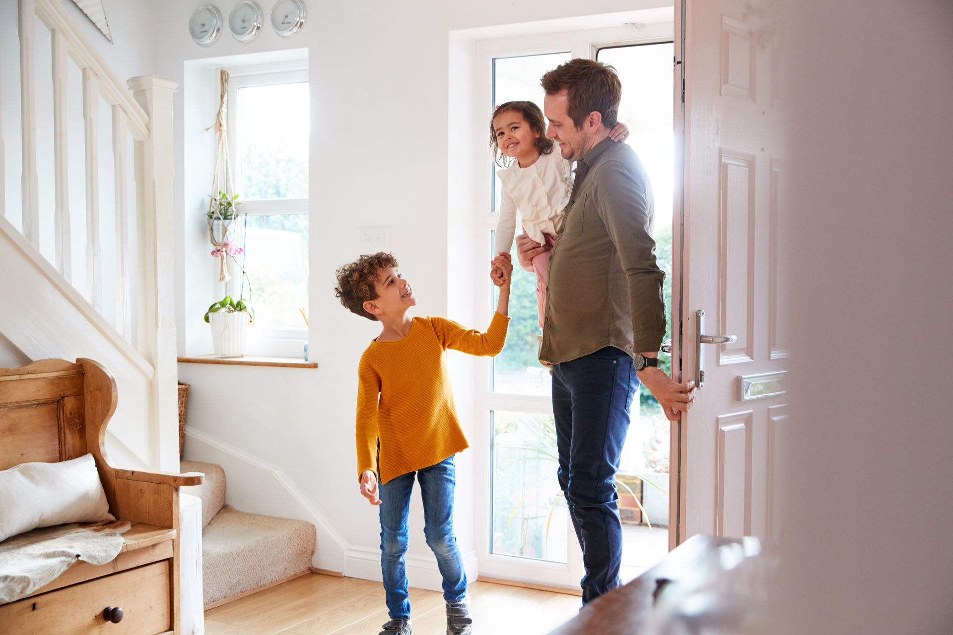 Une famille se salue sur le seuil lumineux d'une maison ; un enfant tend la main vers un homme qui tient un tout-petit.