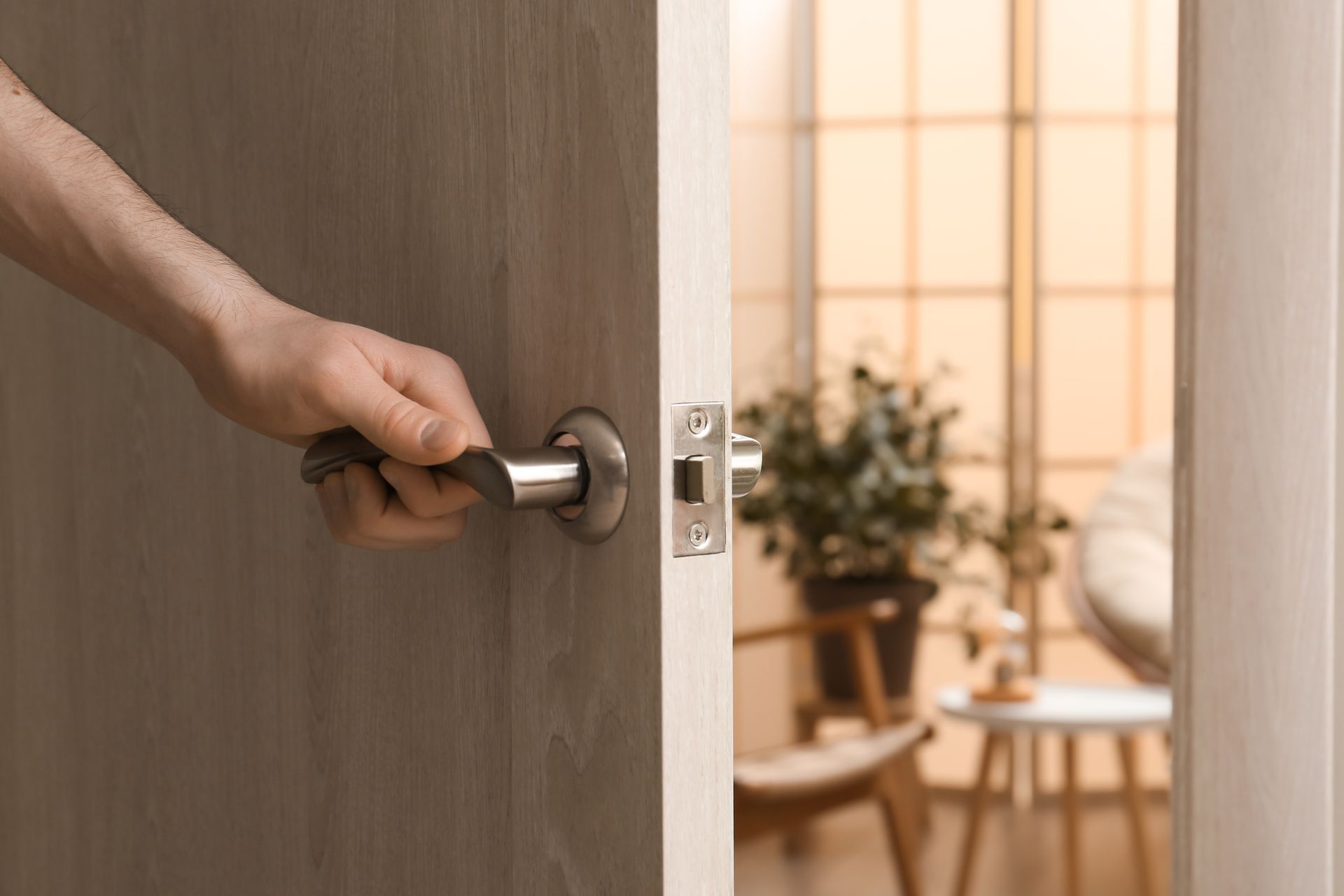 Une main ouvre une porte en bois brun donnant sur une pièce lumineuse avec des plantes et une chaise.