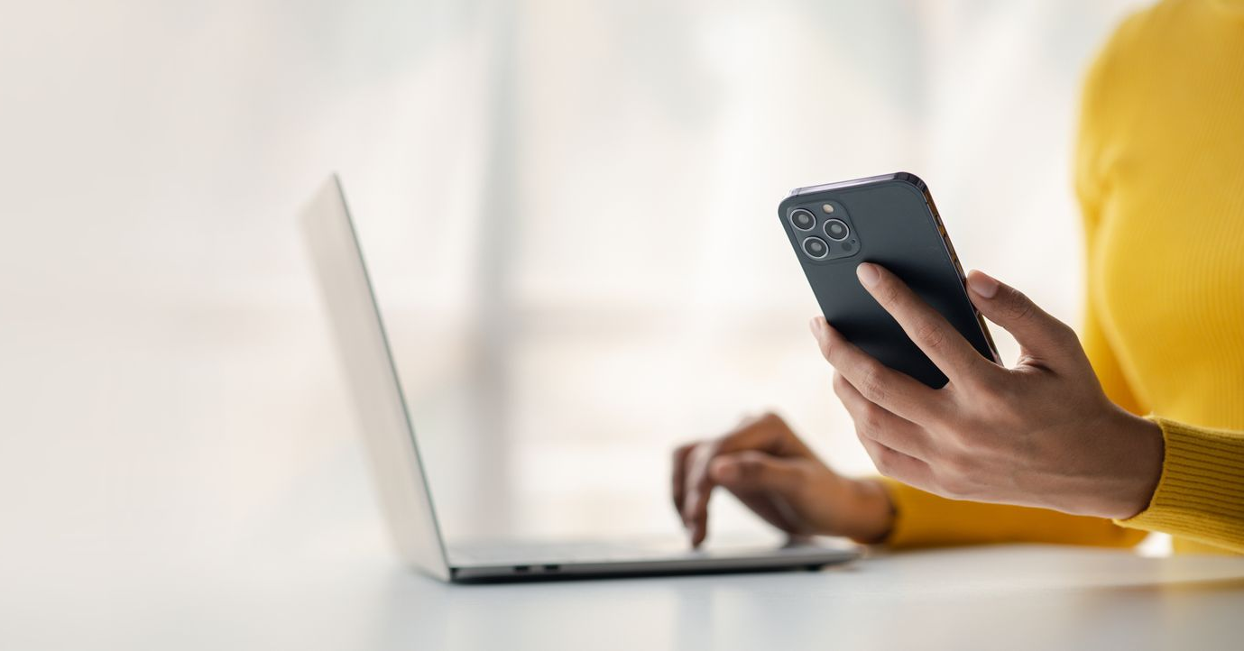 Une personne tenant un smartphone tout en utilisant un ordinateur portable sur un bureau lumineux.