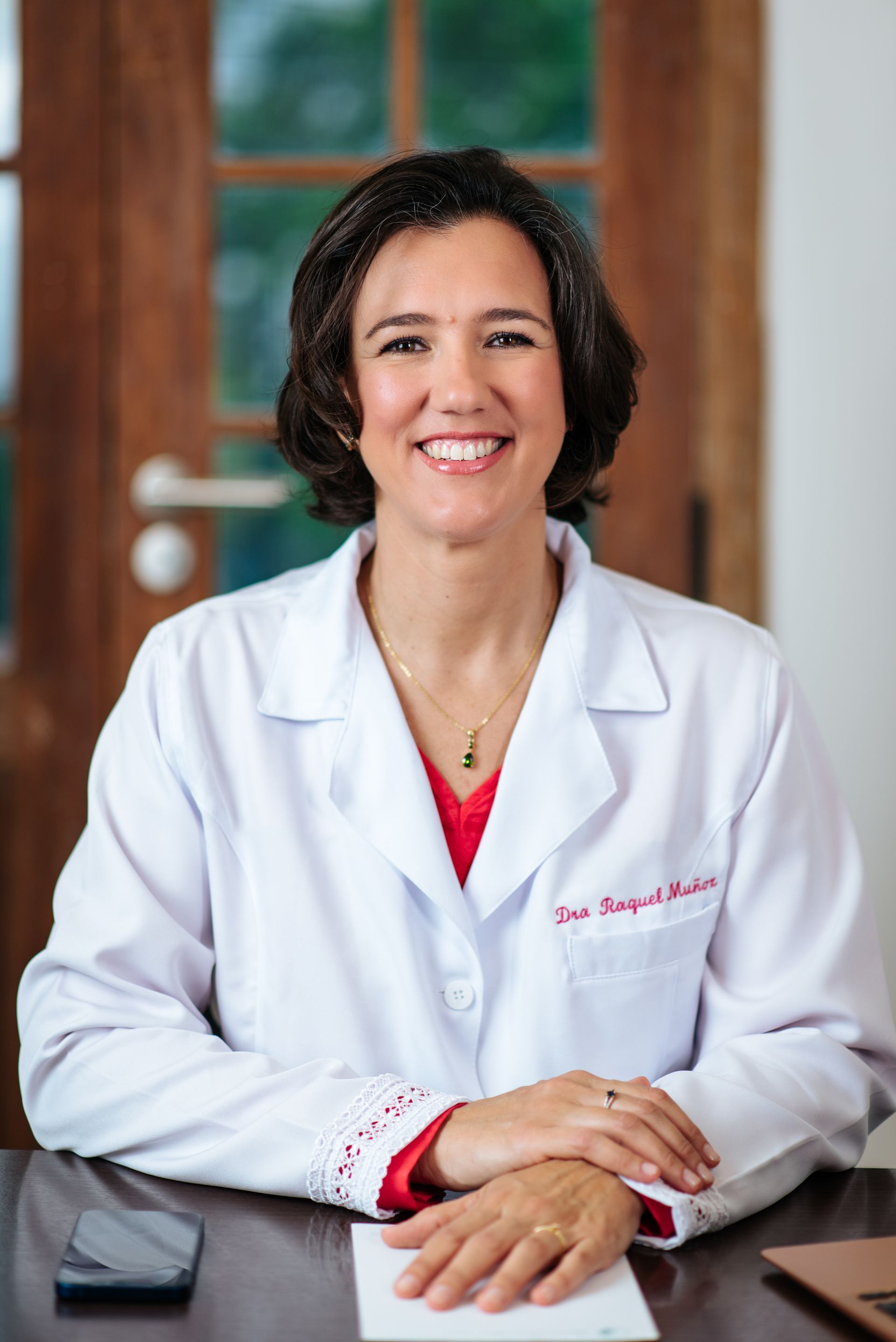Una mujer con una bata de laboratorio está sentada en un escritorio con las manos juntas y sonriendo.