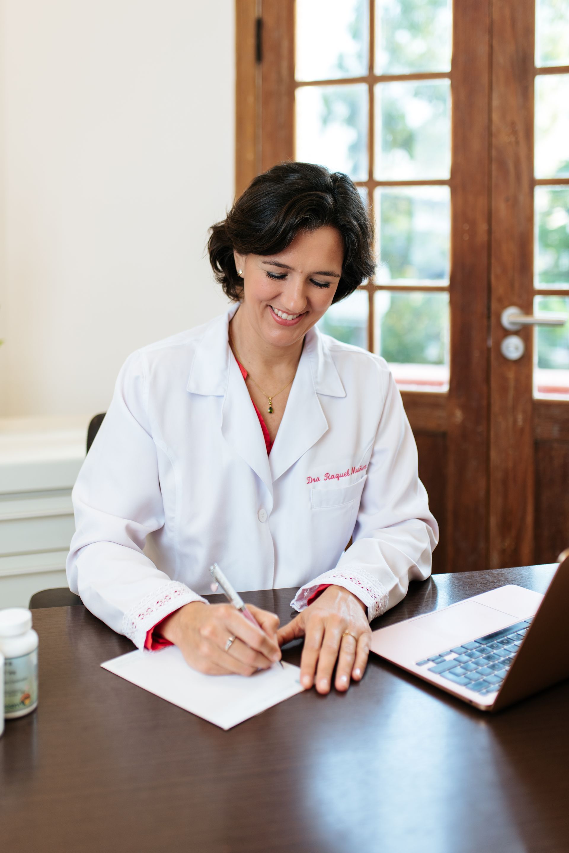 Una mujer con una bata de laboratorio está sentada en un escritorio escribiendo en un trozo de papel.