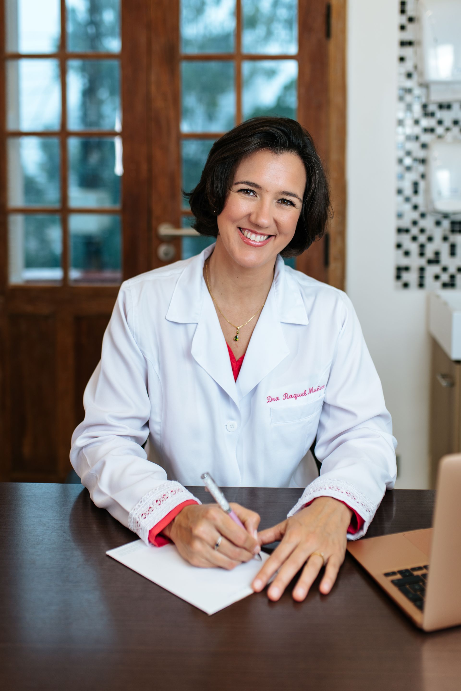Una mujer con una bata de laboratorio está sentada en un escritorio escribiendo en un trozo de papel.