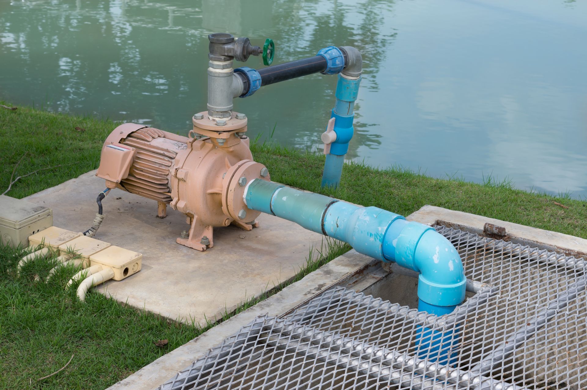Pompe industrielle avec des tuyaux, située près d’un étang et installée sur une dalle de béton, avec une grille métallique à proximité.