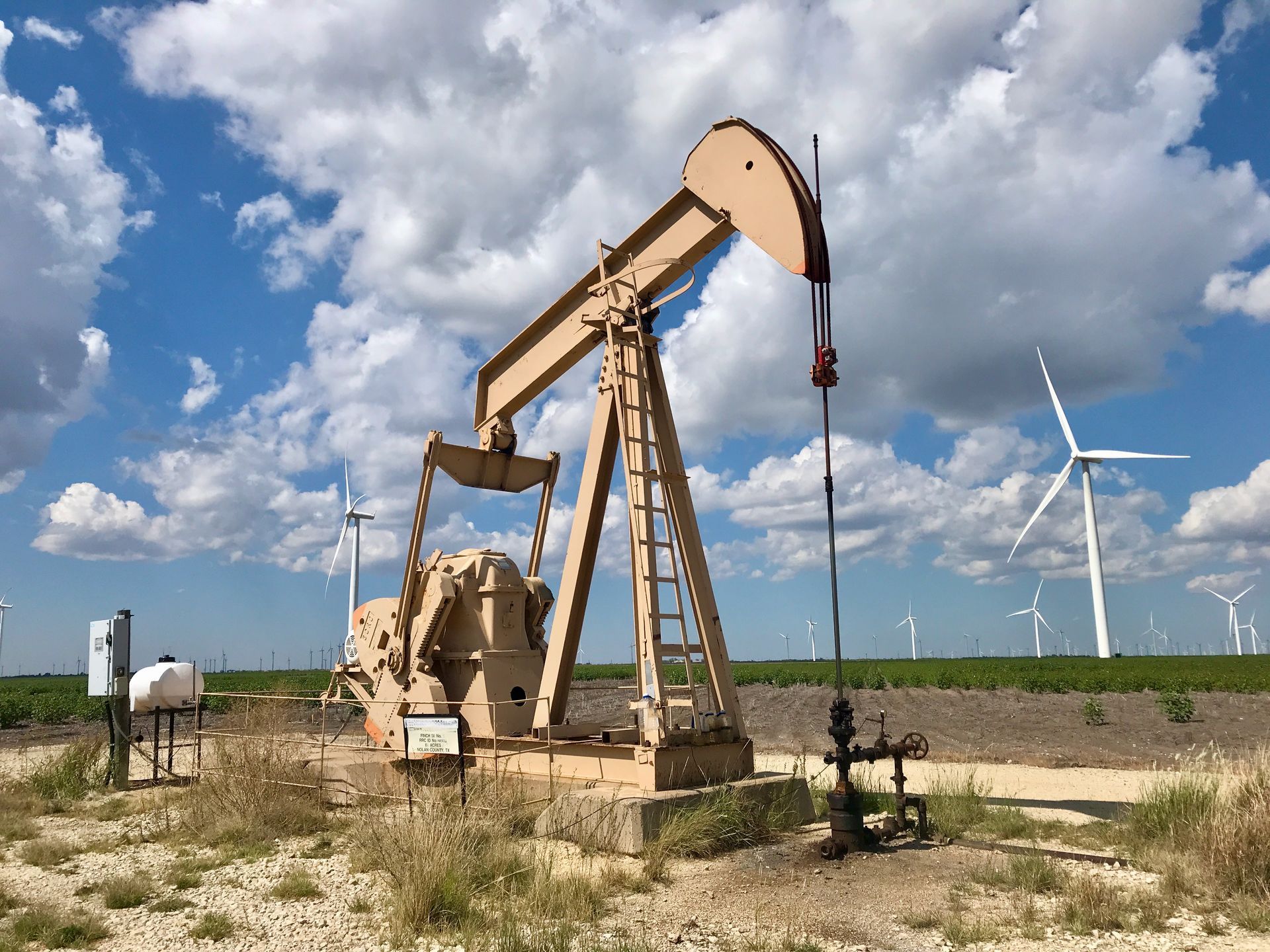 Une pompe à balancier dans un champ rural, avec des éoliennes en arrière-plan.
