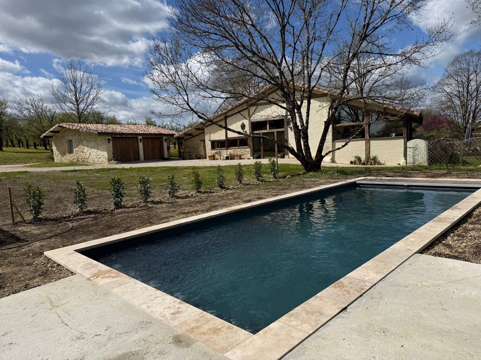 Piscine rectangulaire dans le jardin, à côté d’une maison moderne, avec des arbres dénudés et un ciel nuageux