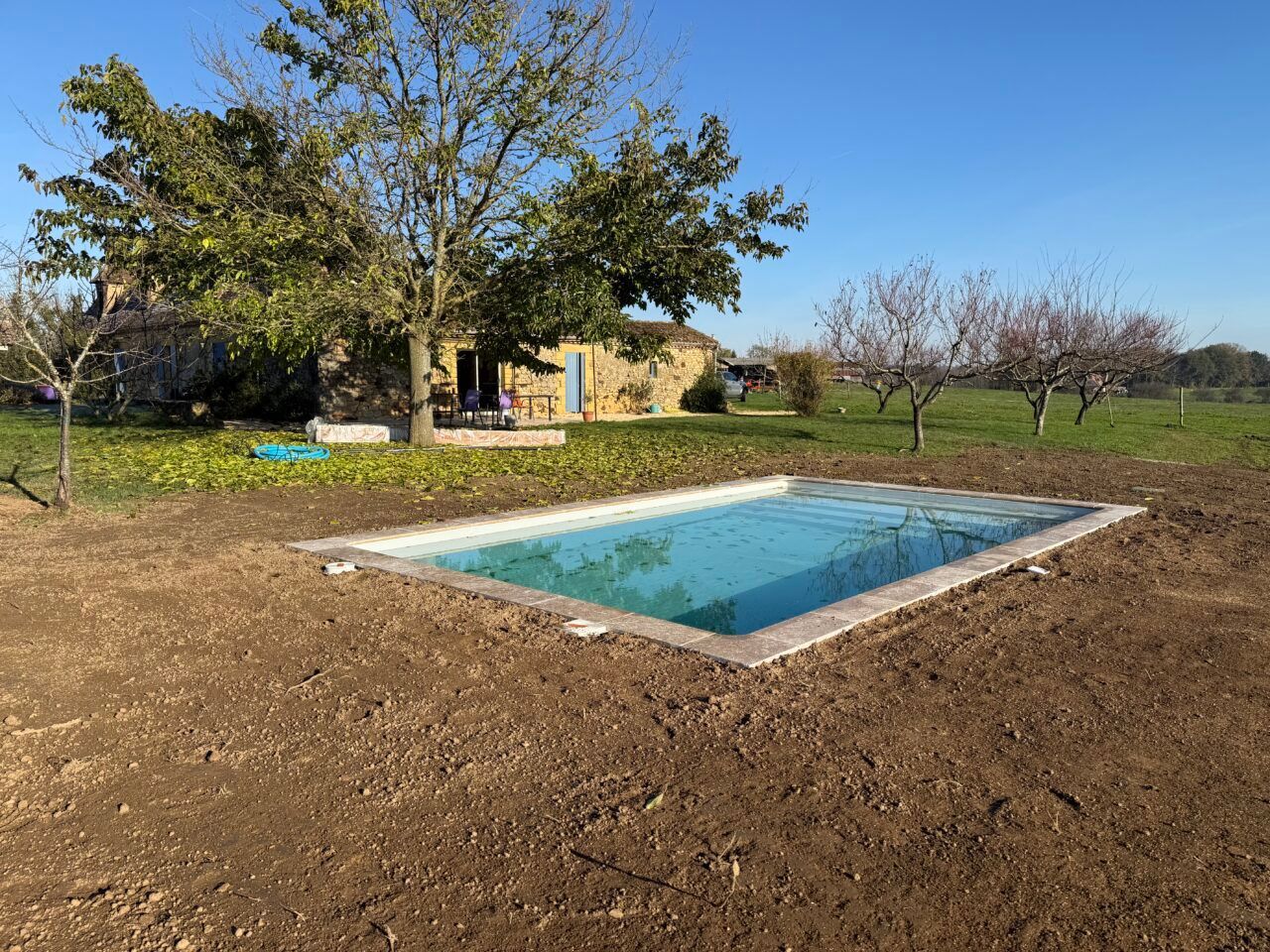 Piscine rectangulaire dans le jardin d’une petite maison, bordée d’arbres et d’herbe sèche, sous un ciel bleu dégagé