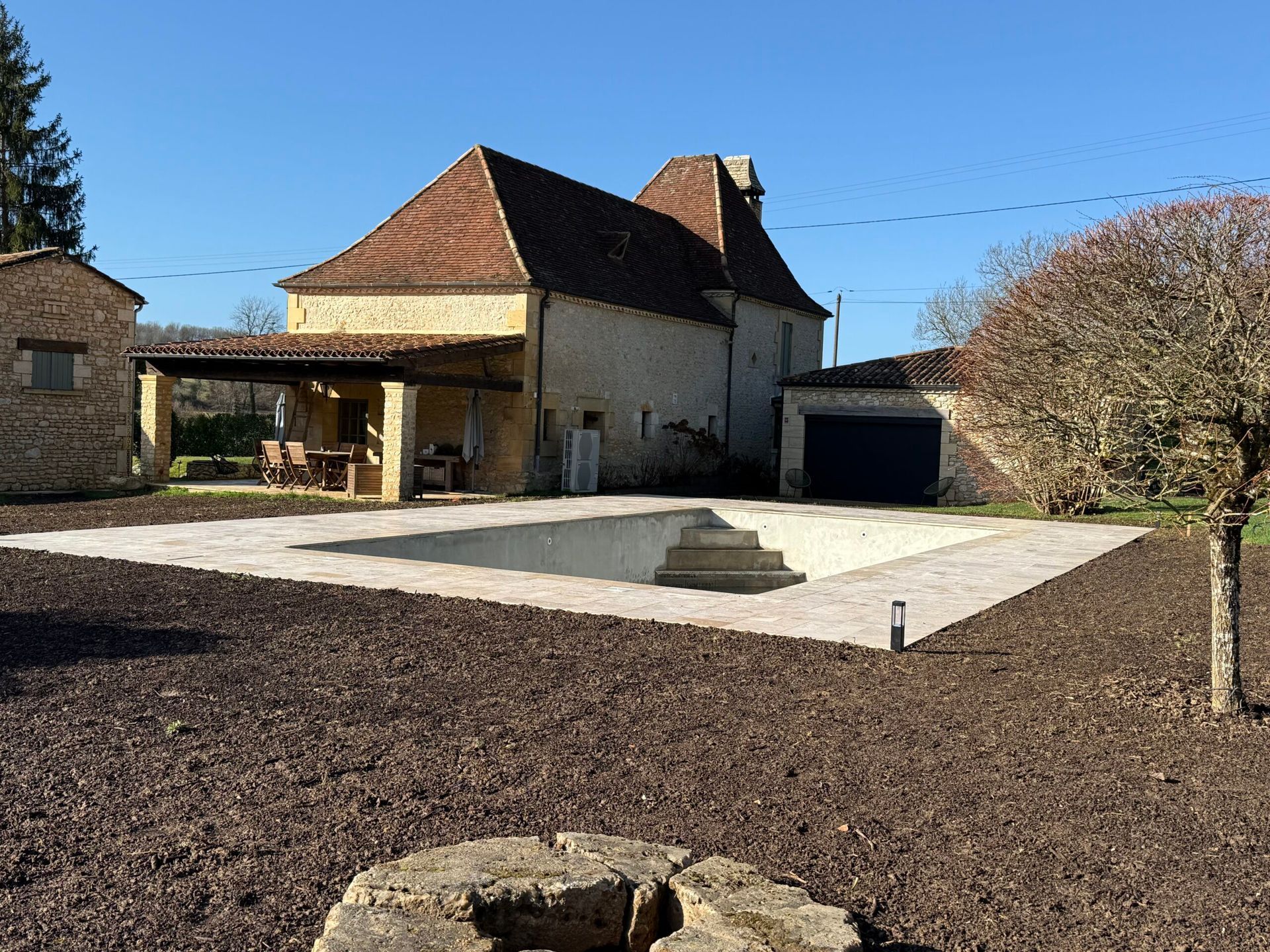 Cour intérieure avec une maison en pierre, une terrasse pavée et des arbres sous un ciel bleu dégagé
