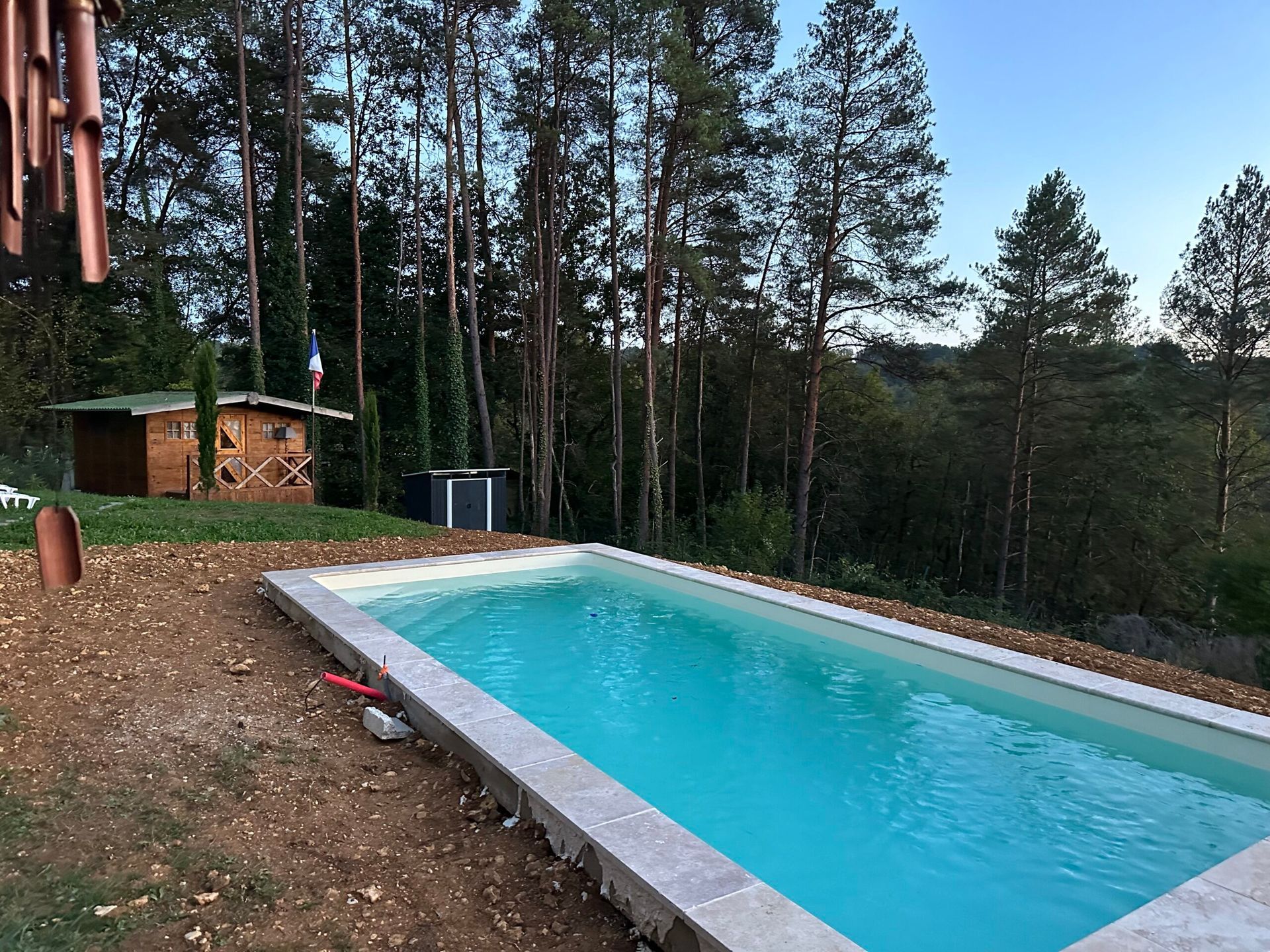 Piscine extérieure rectangulaire à l’eau turquoise, bordée de pins, avec une petite cabane en bois au crépuscule
