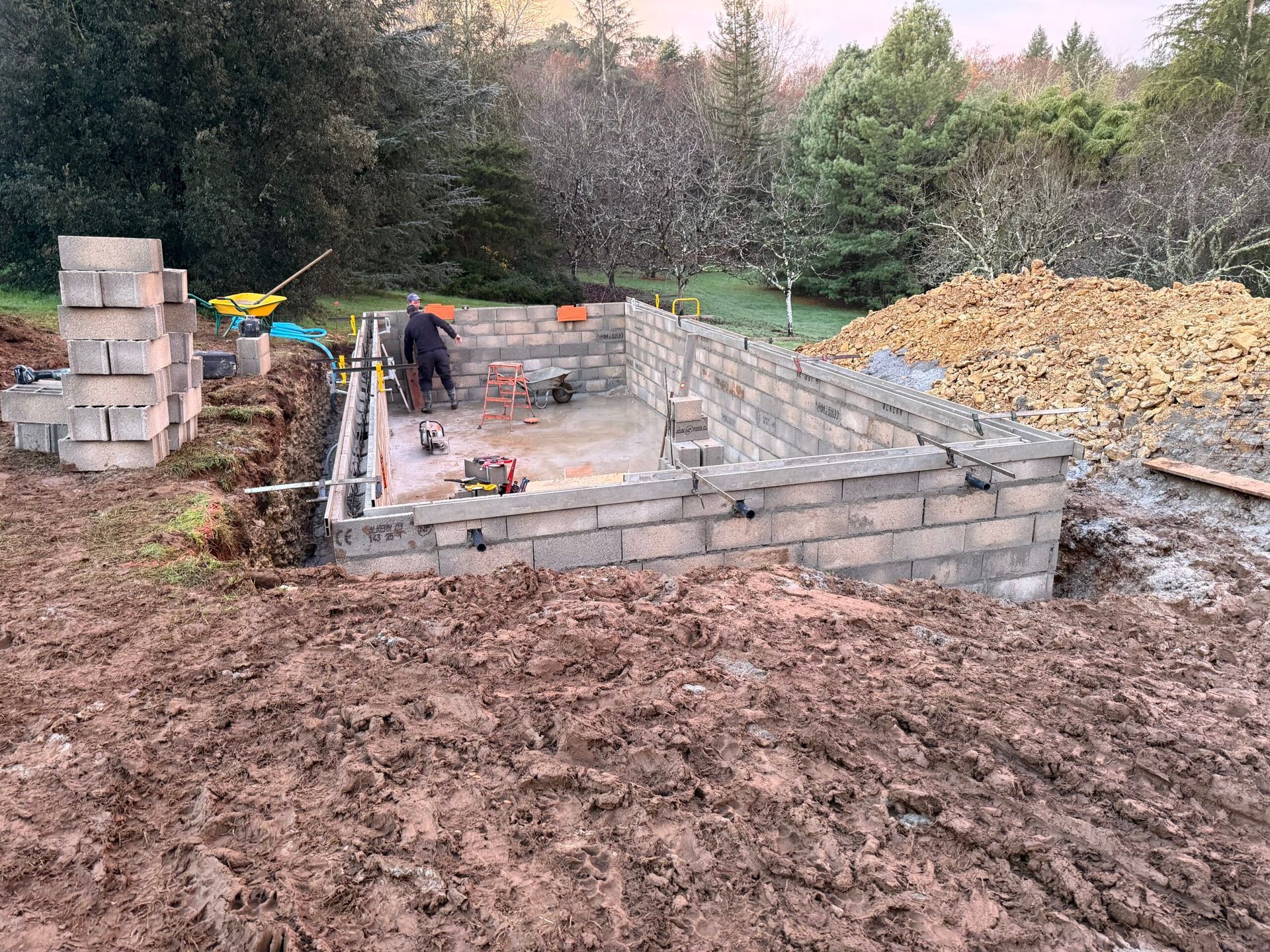 Fondation en parpaings partiellement construite sur un chantier boisé, avec des ouvriers et des tas de terre.