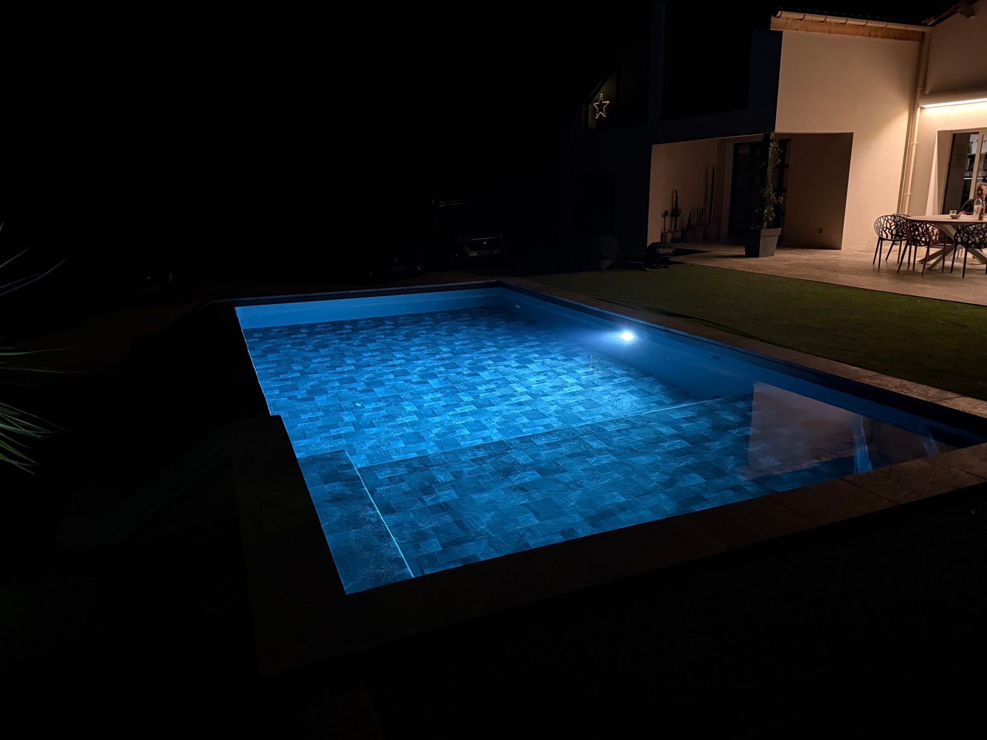 Piscine éclairée en bleu la nuit, à côté d’une maison et de chaises de terrasse