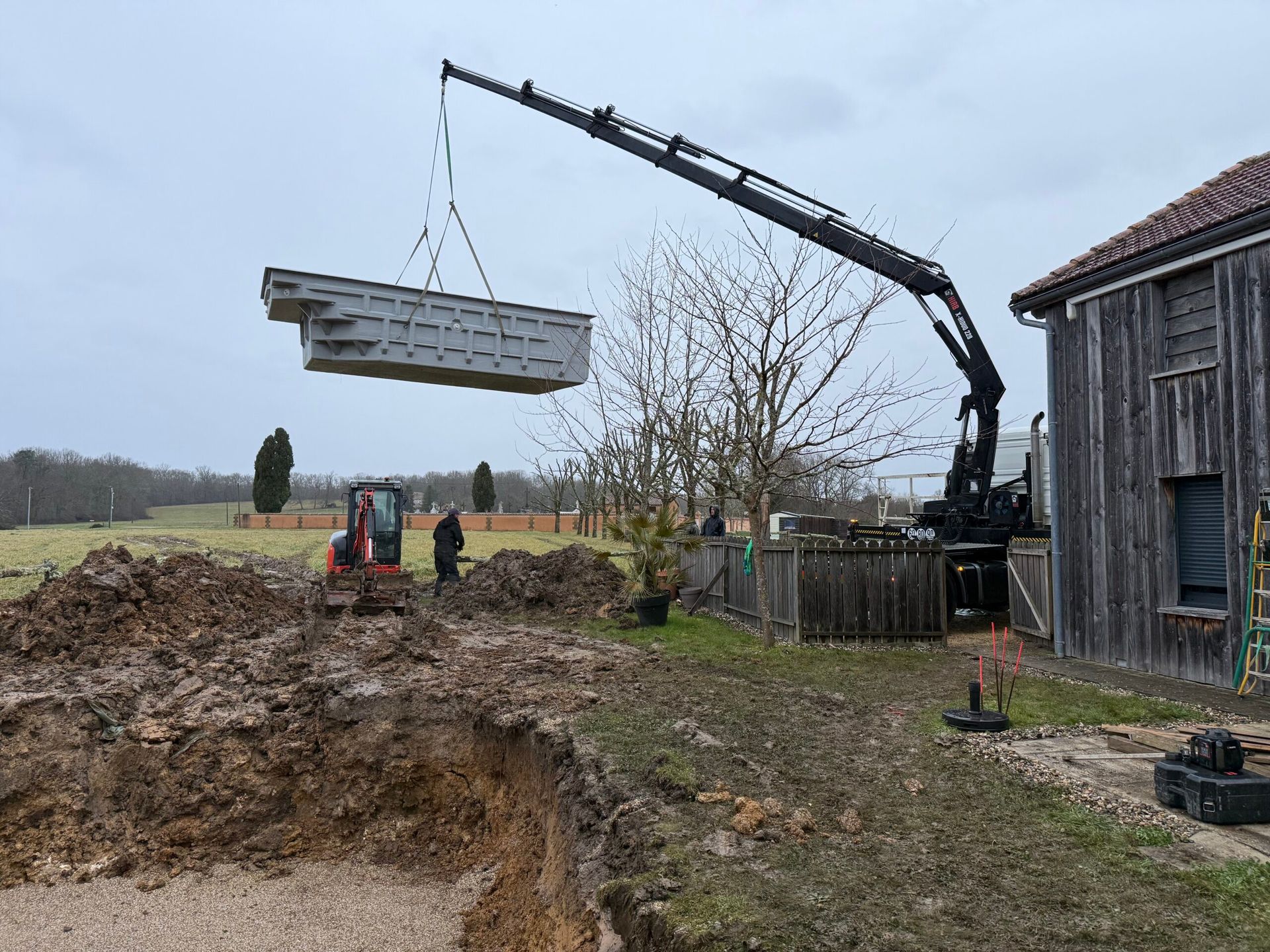 Grue soulevant une grosse poutre métallique à côté d’une maison à la campagne et d’un terrain excavé