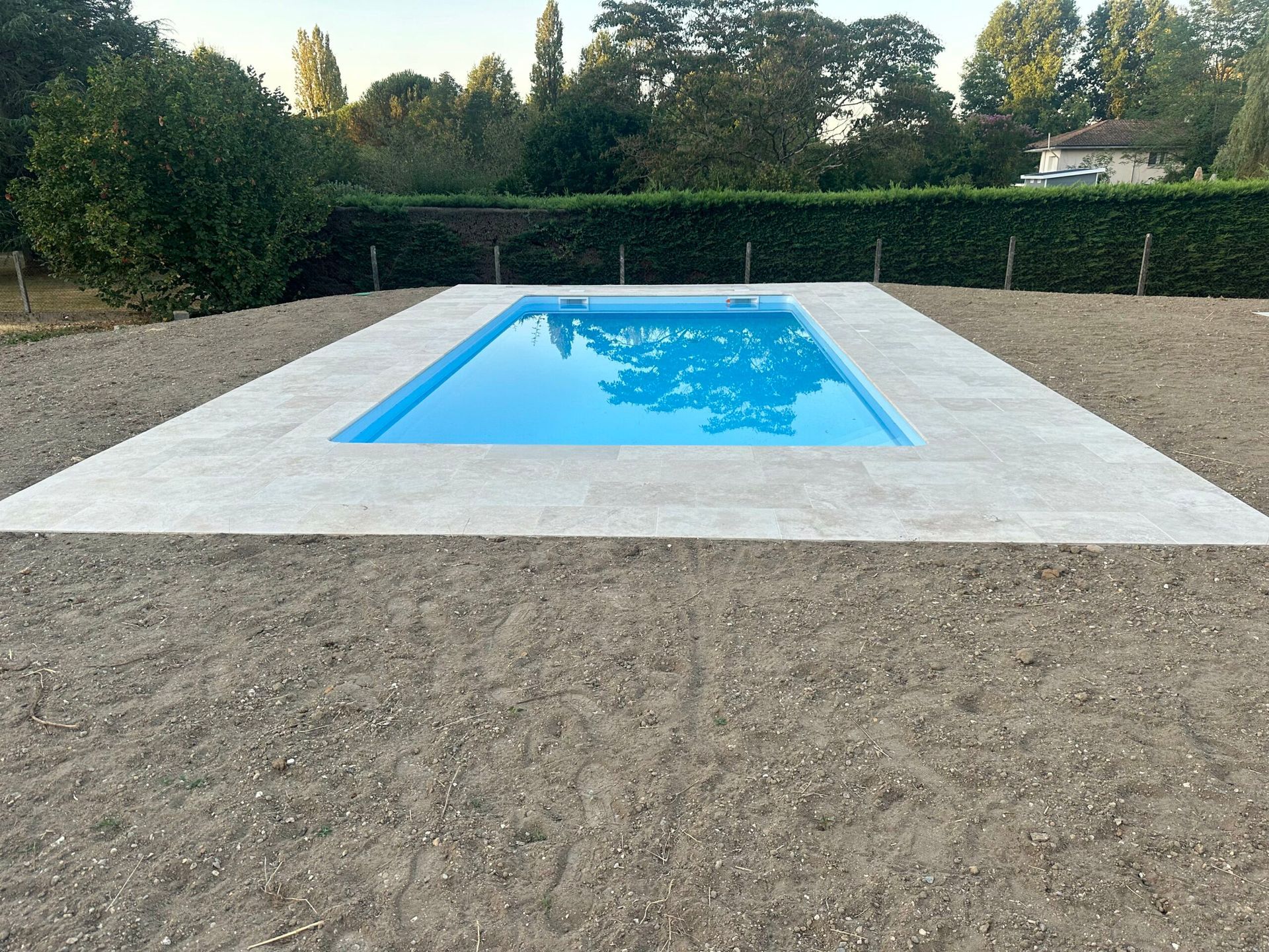 Piscine rectangulaire bleue installée dans une cour gravillonnée, bordée de haies et d’arbres sous un ciel dégagé