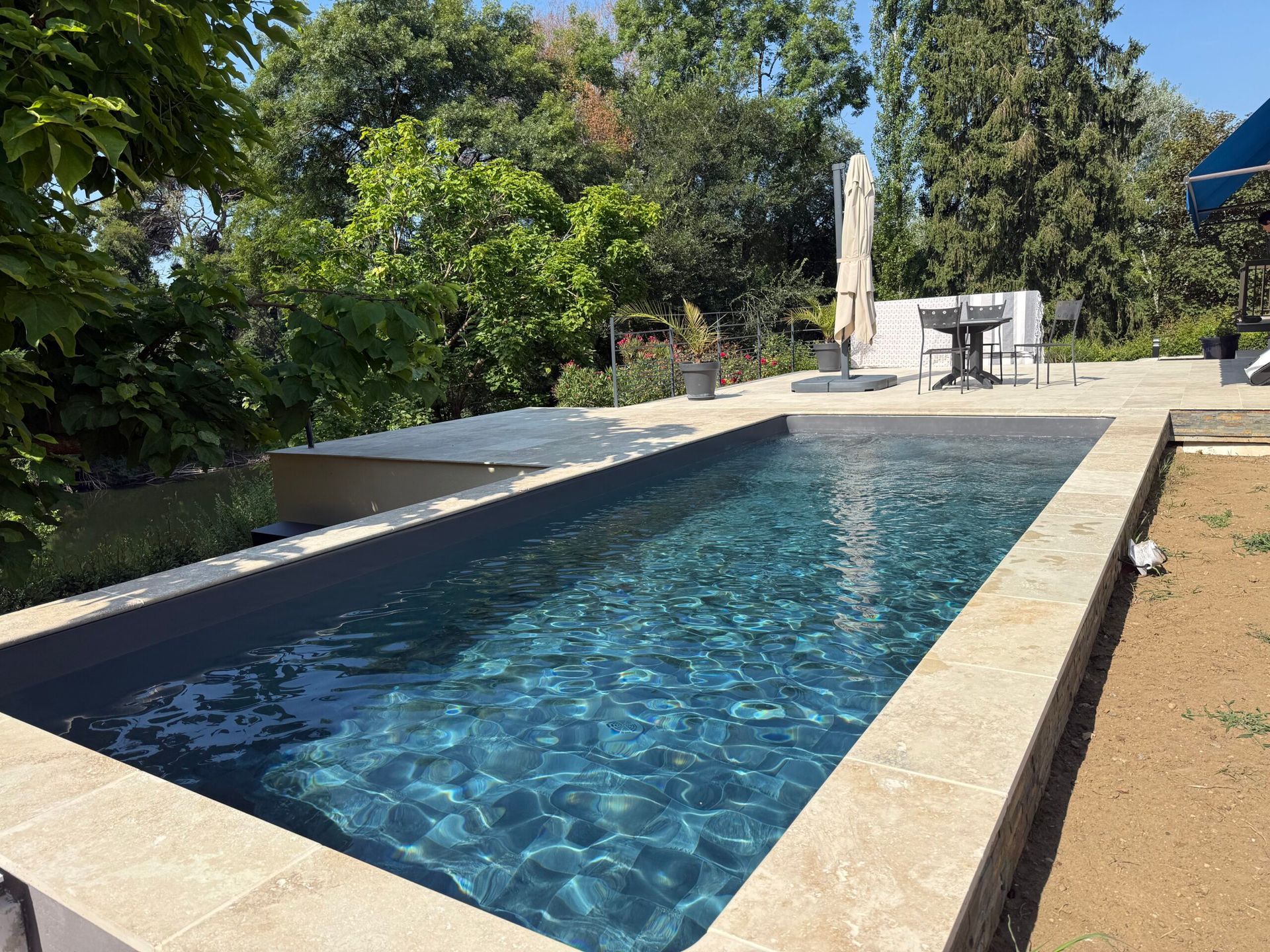 Piscine rectangulaire dans un jardin, à l’eau bleue, entourée d’une terrasse et d’arbres sous un ciel dégagé