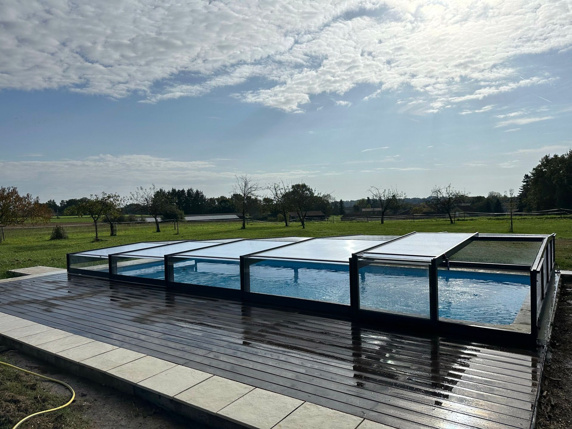 Piscine extérieure rectangulaire sous un ciel nuageux, avec de l’eau bleue et une terrasse sombre
