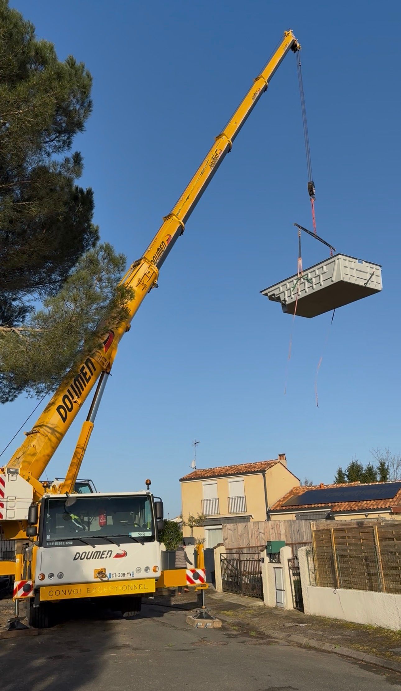 Une grue jaune soulève une grande piscine coque sur un chantier, sous un ciel bleu dégagé