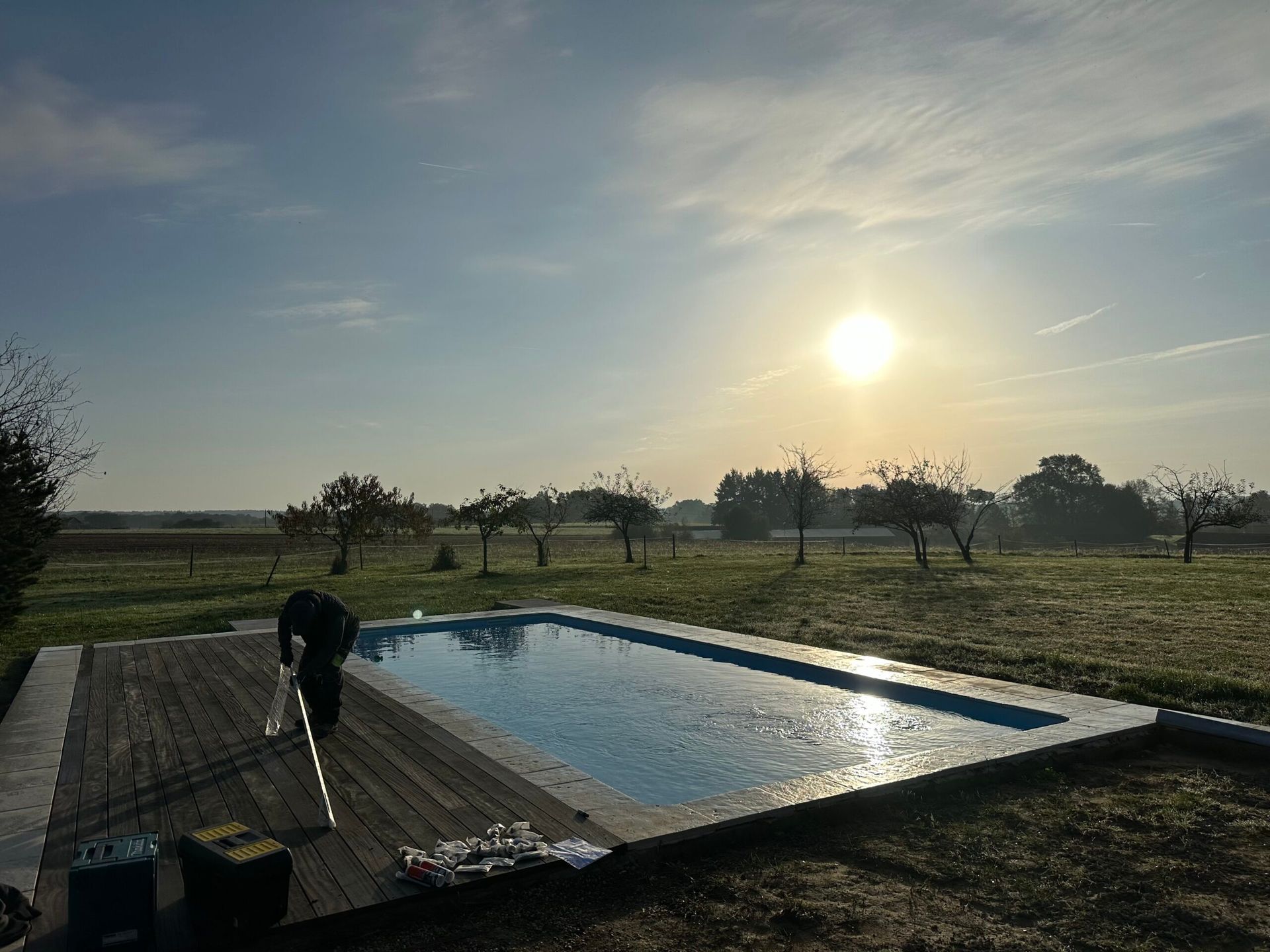 Terrasse au bord d’une piscine, près d’un champ enherbé au lever du soleil, avec une personne penchée sur la terrasse.