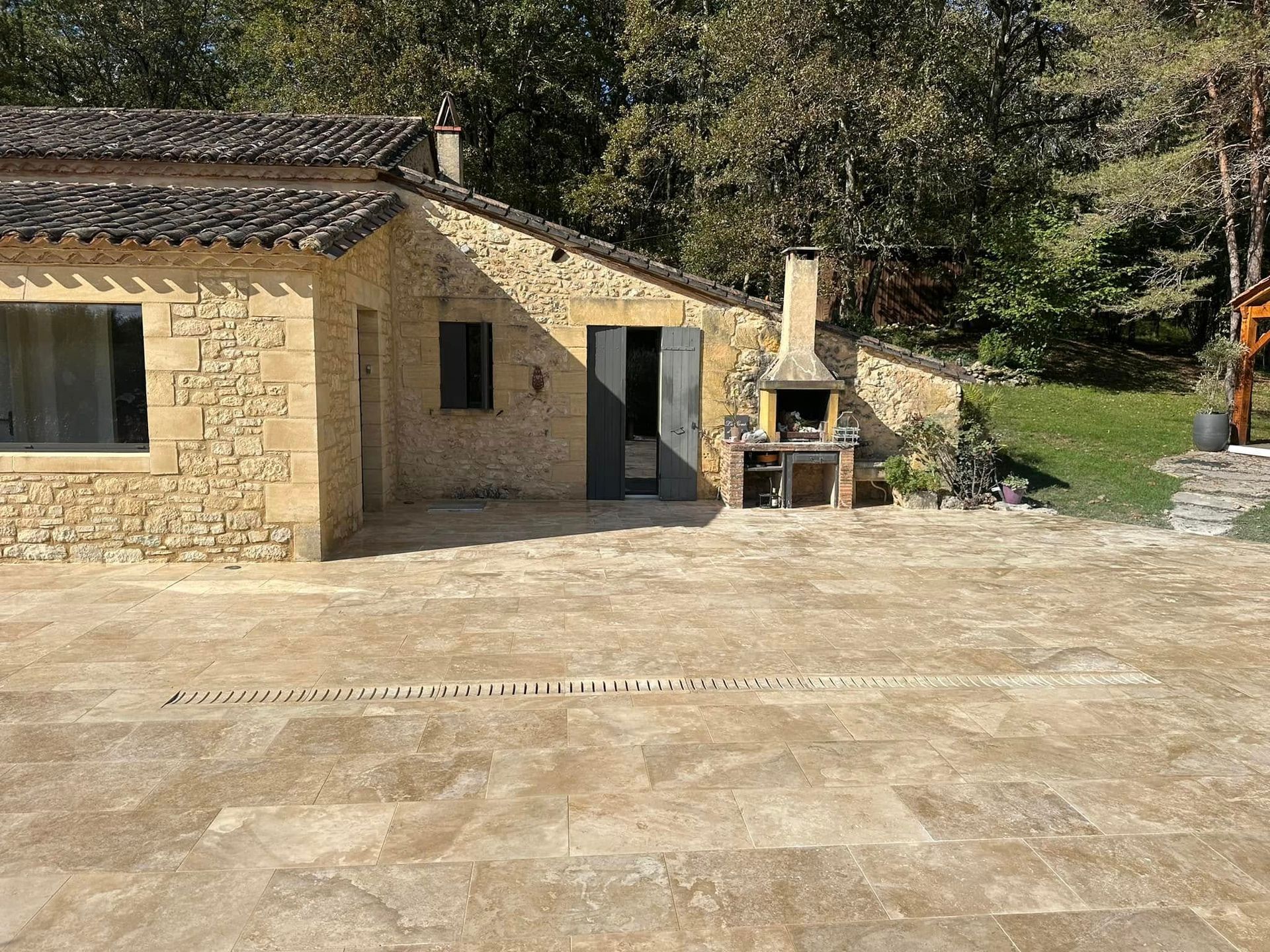 Maison en pierre avec toit de tuiles et barbecue extérieur dans une cour en terre ensoleillée, bordée d’arbres