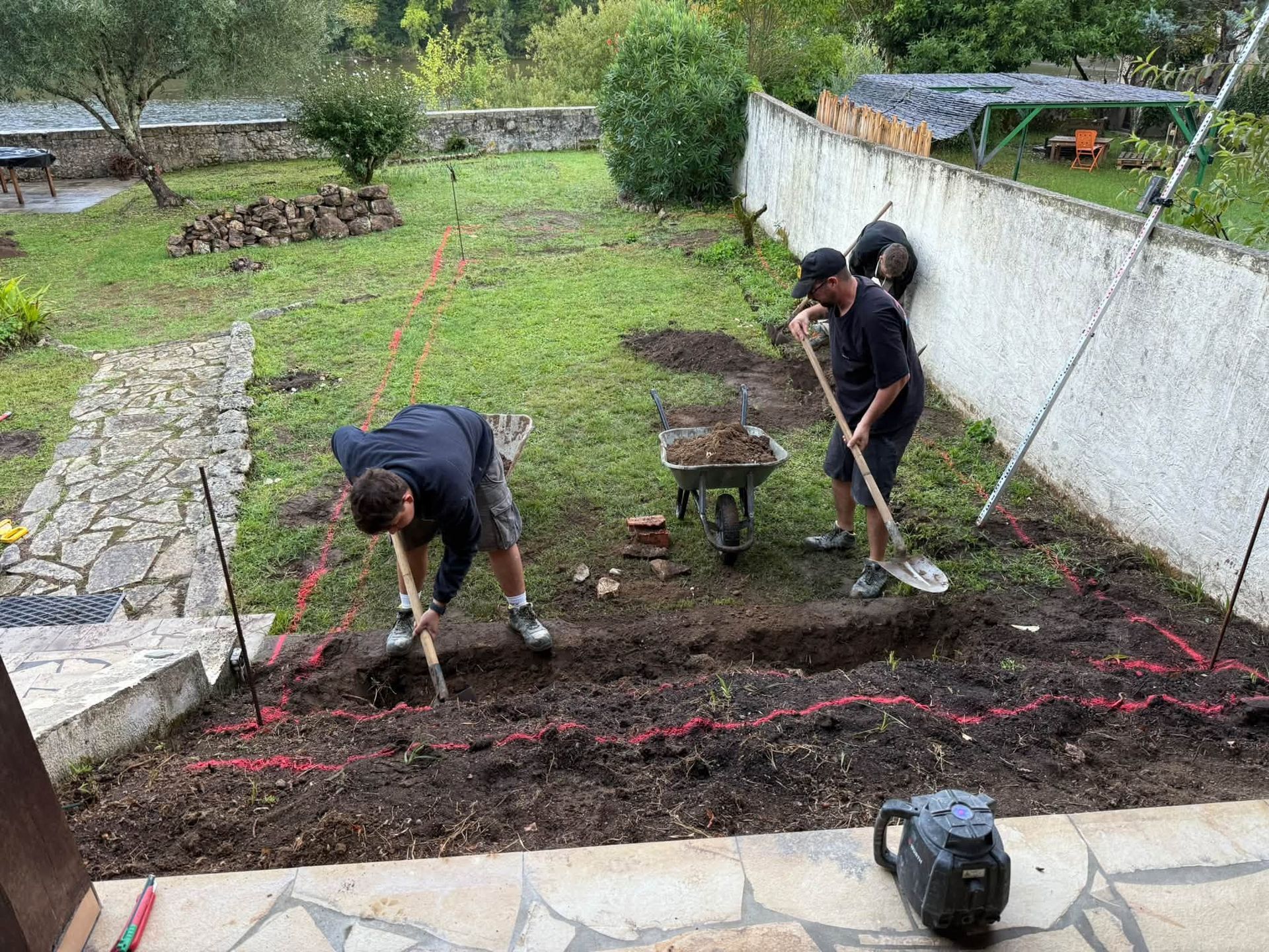 Deux personnes bêchent un parterre de jardin à la pelle, près d’un mur blanc et d’une allée en pierre.