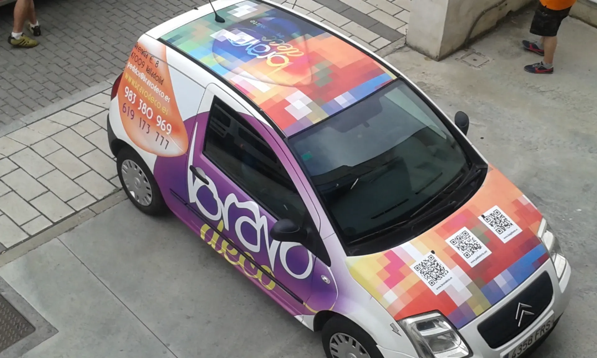 Coche con publicidad colorida, aparcado sobre hormigón. Tres códigos QR en el capó.