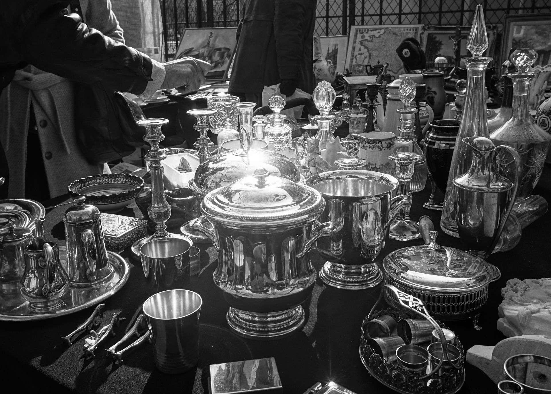 Service à thé en argent assorti et vaisselle sur une table encombrée, avec une personne qui tend la main en arrière-plan.