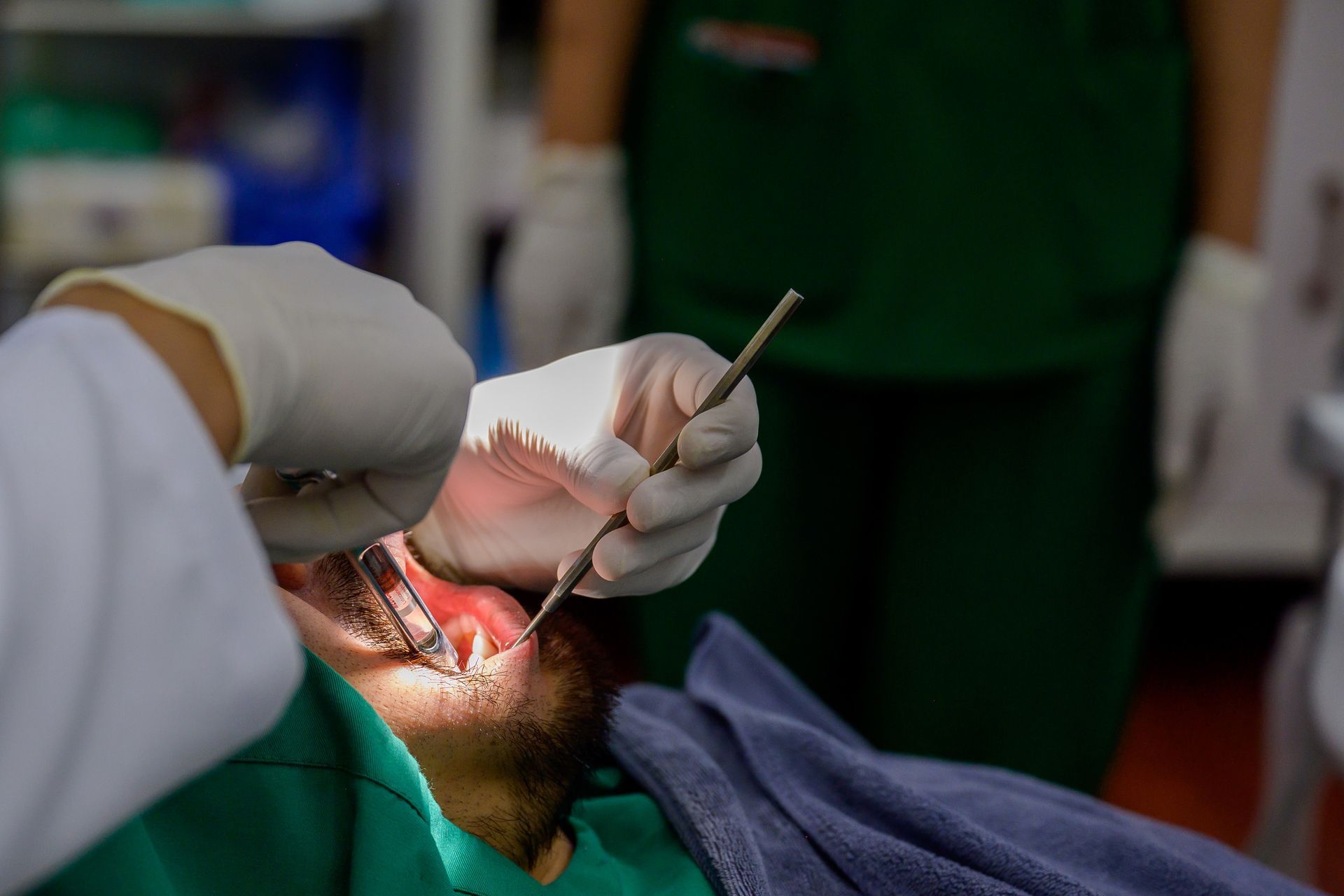 Un dentista examina la boca de un paciente con un instrumento. El paciente lleva una bata quirúrgica verde.