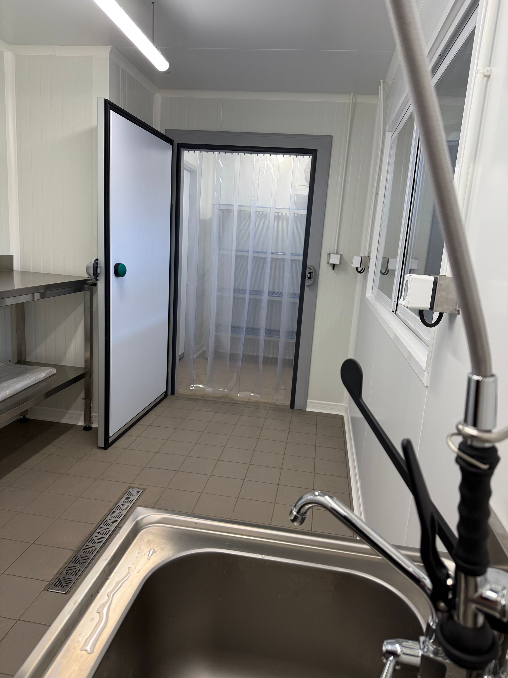 Une cuisine professionnelle avec un évier en métal au premier plan, une porte avec des rideaux en plastique et des murs blancs.