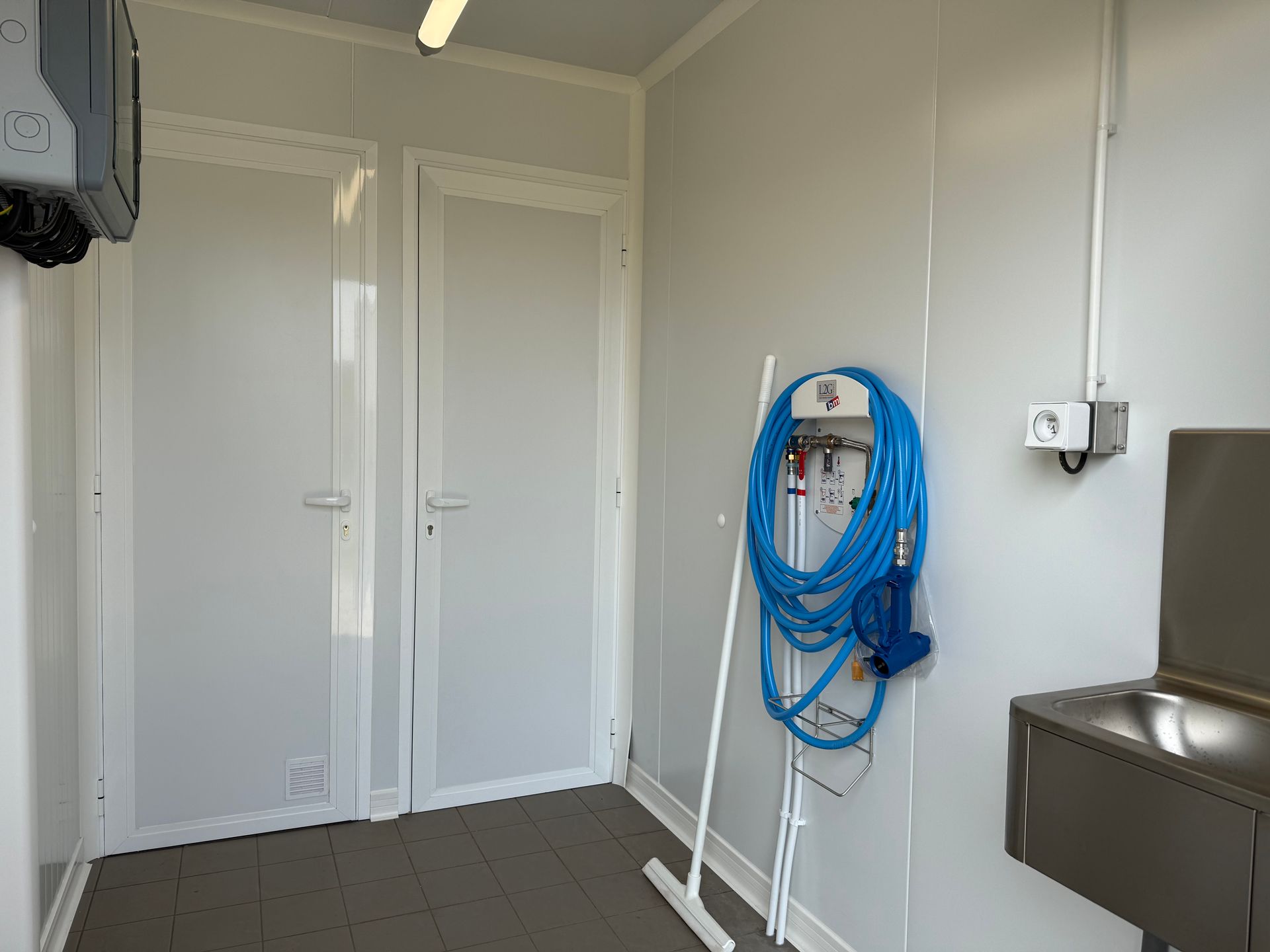 Une pièce avec deux portes blanches, un tuyau d'arrosage bleu enroulé au mur, une serpillière et un évier utilitaire en acier inoxydable.