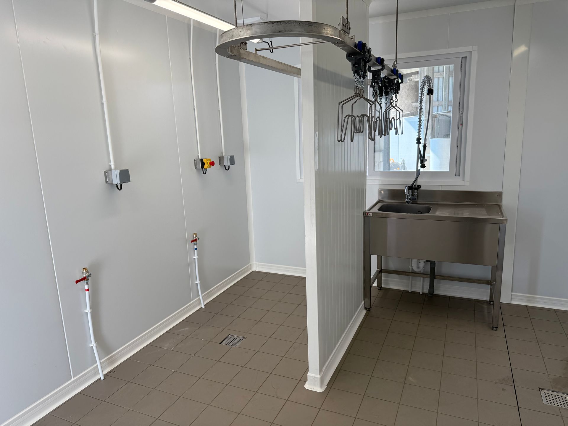 Une salle de transformation alimentaire stérile avec des murs blancs, un sol carrelé, un évier en acier inoxydable et des rails de suspension pour la viande.