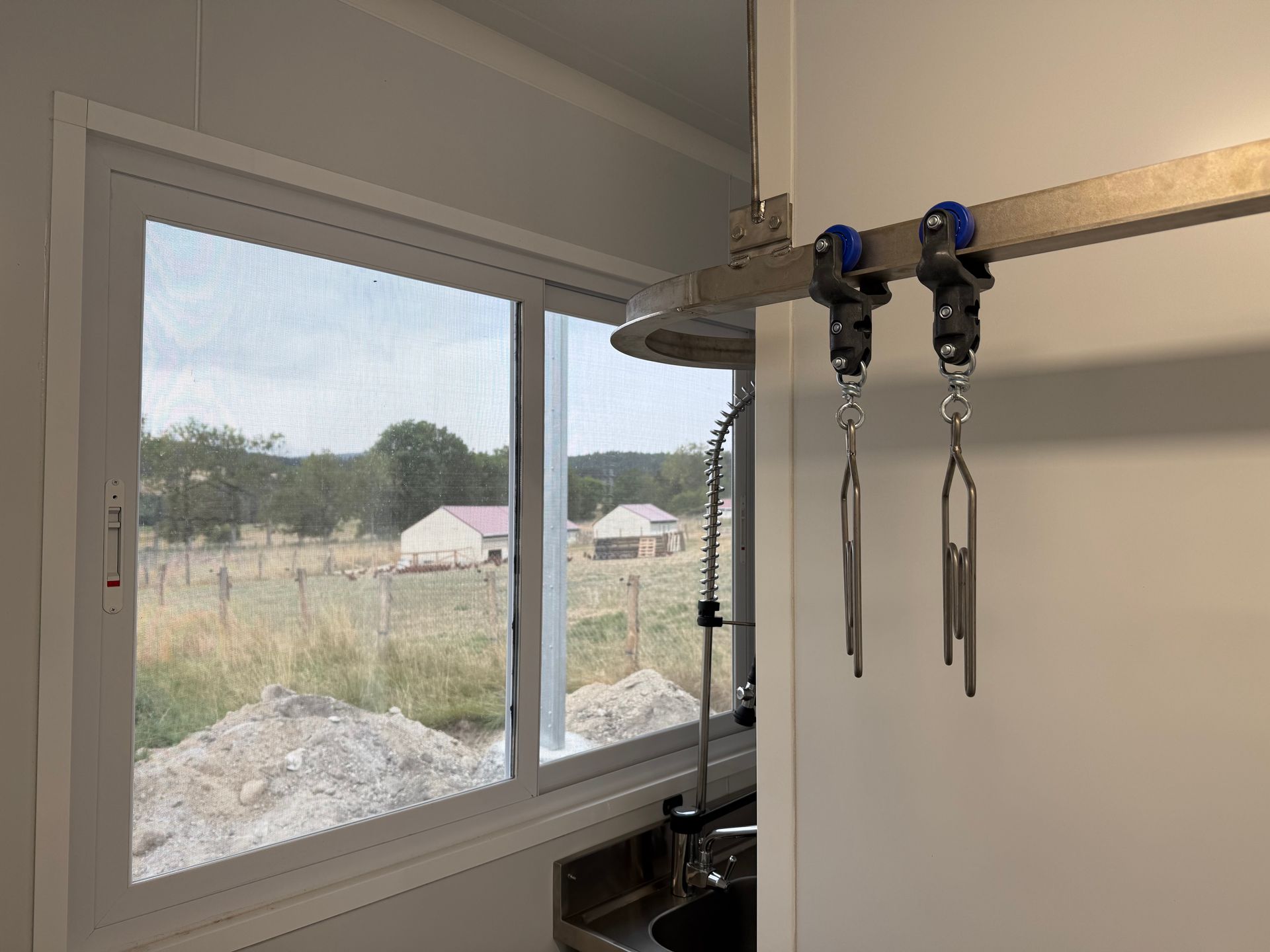 Vue intérieure, à travers une fenêtre, d'un paysage rural avec une voie ferrée métallique et deux crochets de transformation de viande suspendus au premier plan.