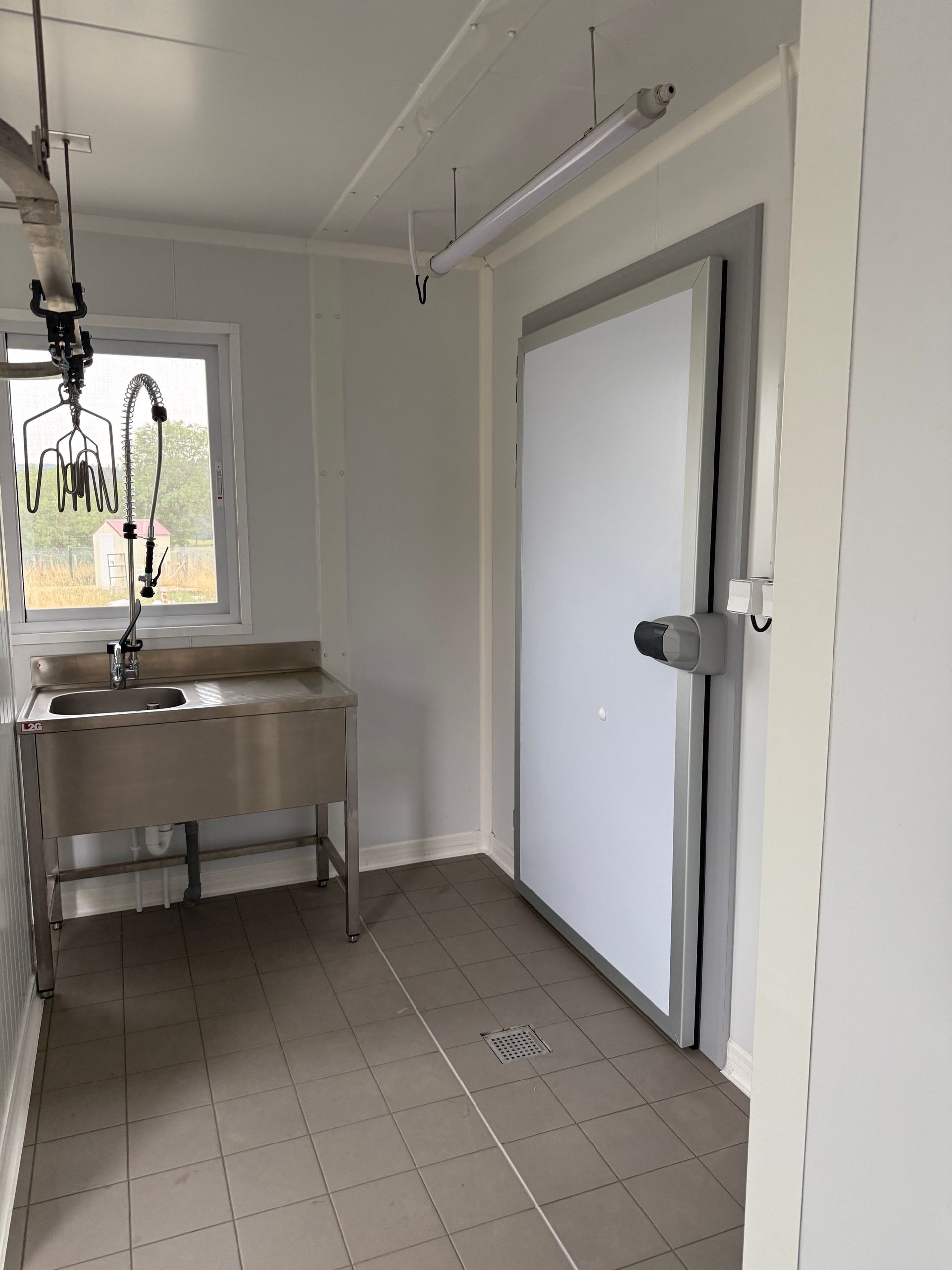Vue intérieure d'une pièce avec un évier en acier inoxydable, une porte de chambre froide et un sol carrelé avec une bonde.
