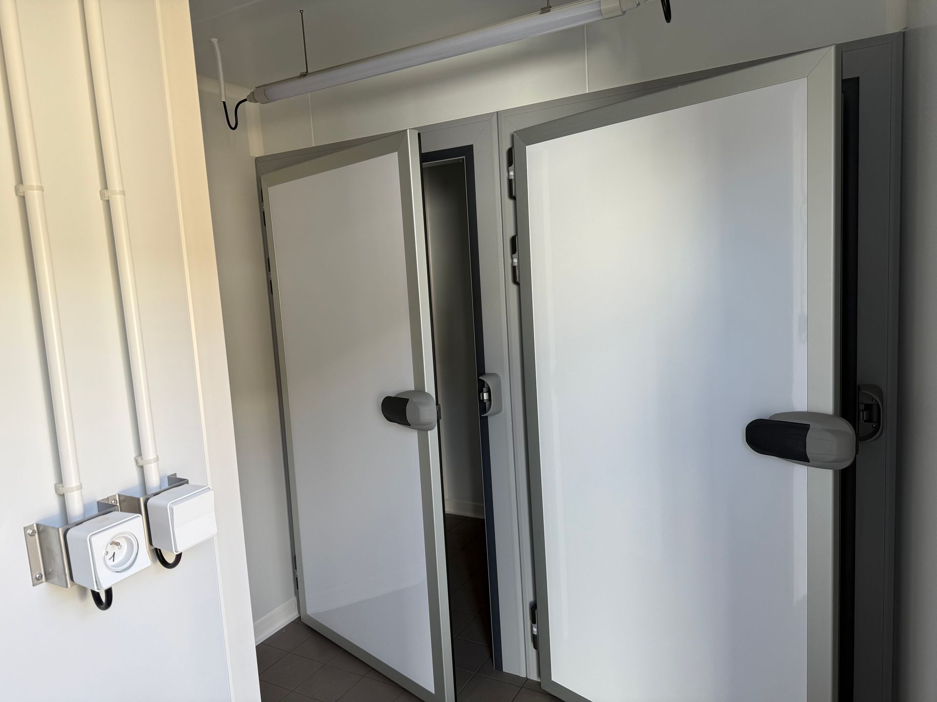Deux portes blanches industrielles de chambre froide ou de congélateur encastrées dans un mur simple, avec un petit boîtier utilitaire monté sur la gauche.