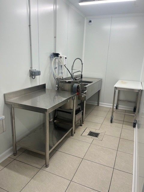 Des tables de préparation en acier inoxydable et un évier commercial avec robinet à douchette dans une buanderie ou une cuisine lumineuse aux murs blancs.