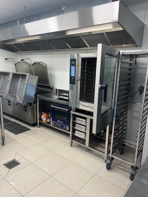 Une cuisine commerciale en acier inoxydable comprenant un grand four, un support à plateaux, une hotte aspirante et du matériel de préparation.