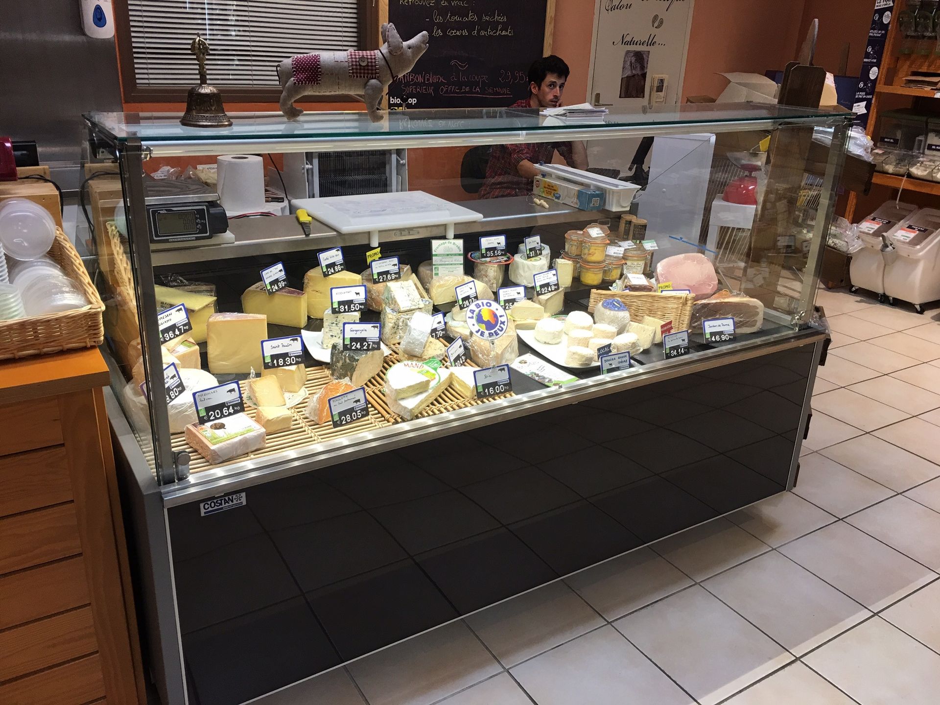 Une vitrine à fromages bien garnie dans un magasin, avec une variété de fromages, une petite figurine de cochon et une cloche sur le dessus.