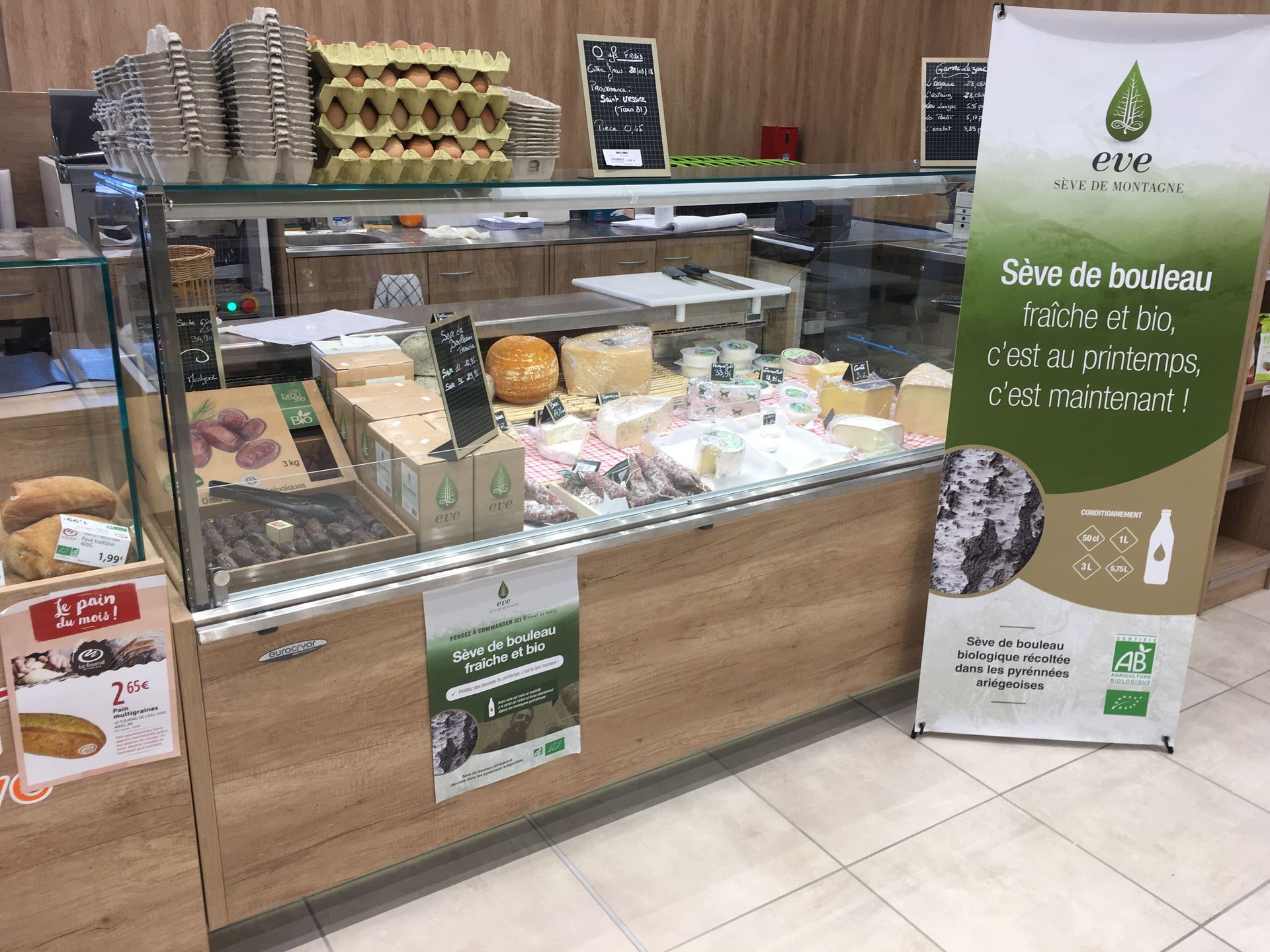 Une vitrine réfrigérée de supermarché remplie de fromages et d'œufs, à côté d'une grande banderole publicitaire pour la sève de bouleau.